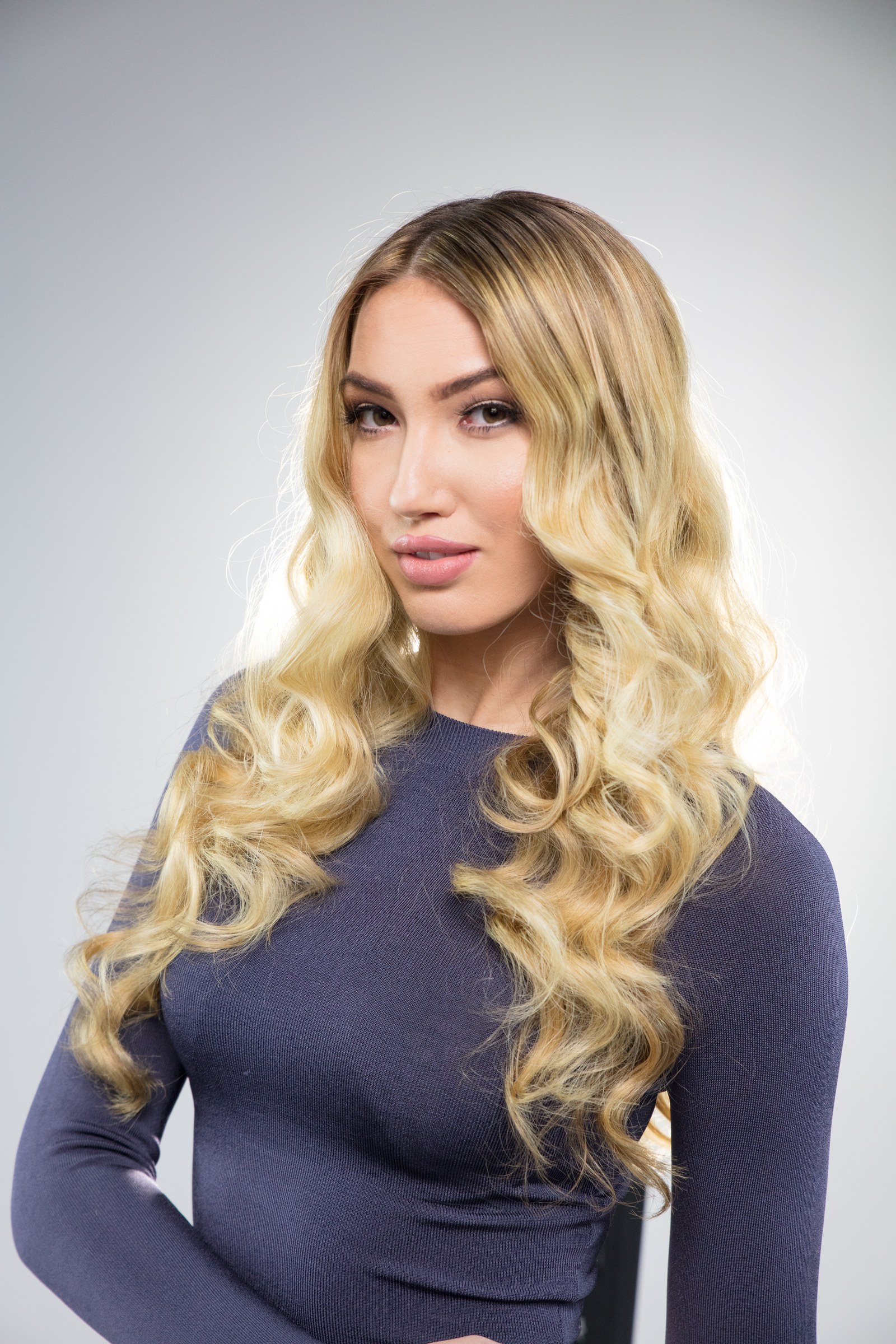 Blonde model with long curled hair wearing a dark top in a studio portrait.