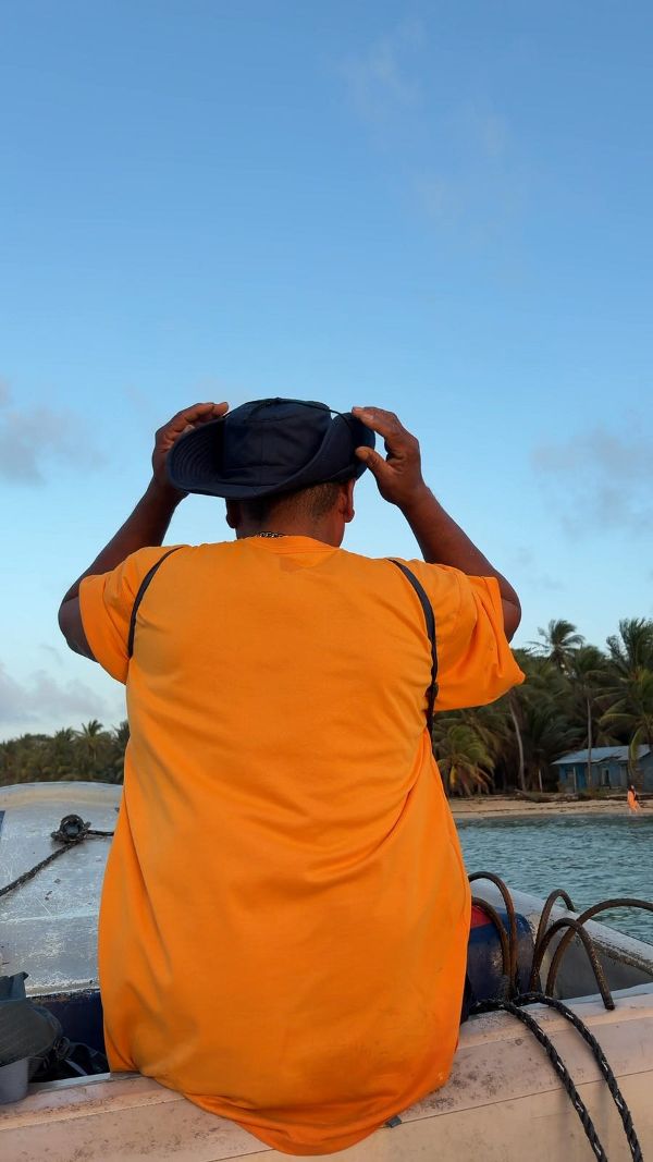 a figure in an orange shirt seen from behind on a boat, hands raised to a hat, water and palms ahead.