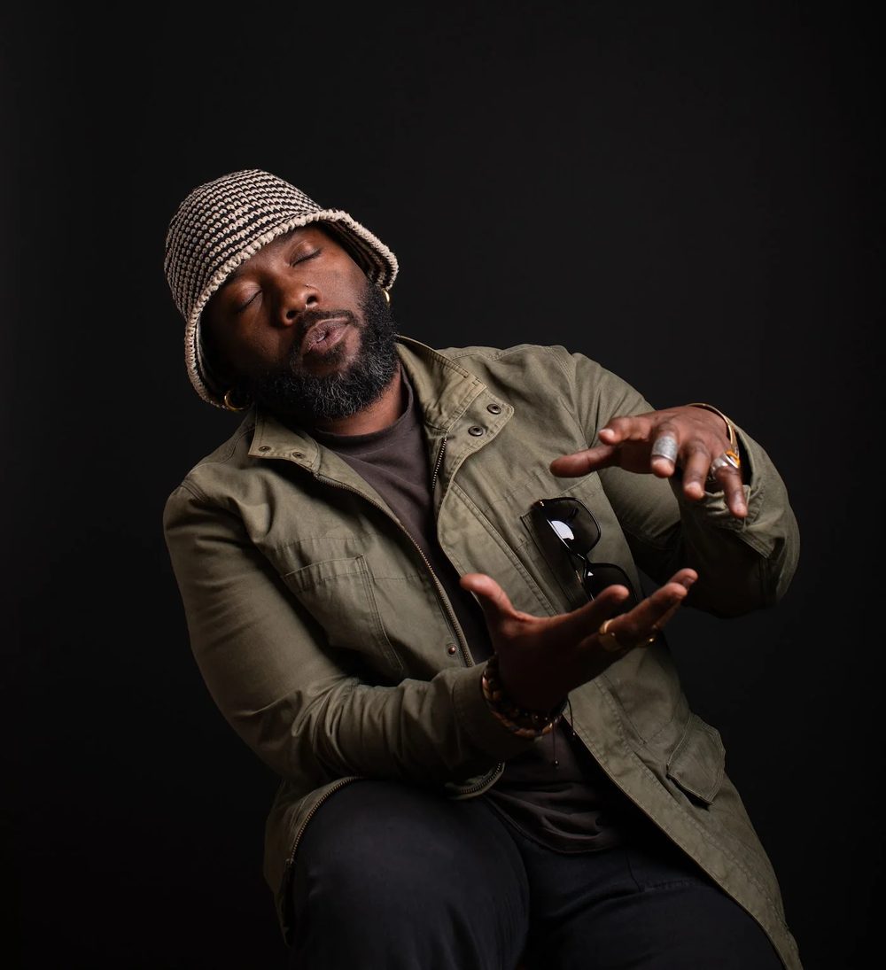 quincéy in a striped crochet hat and green jacket, eyes closed, head tilted back, both hands shaping the air.