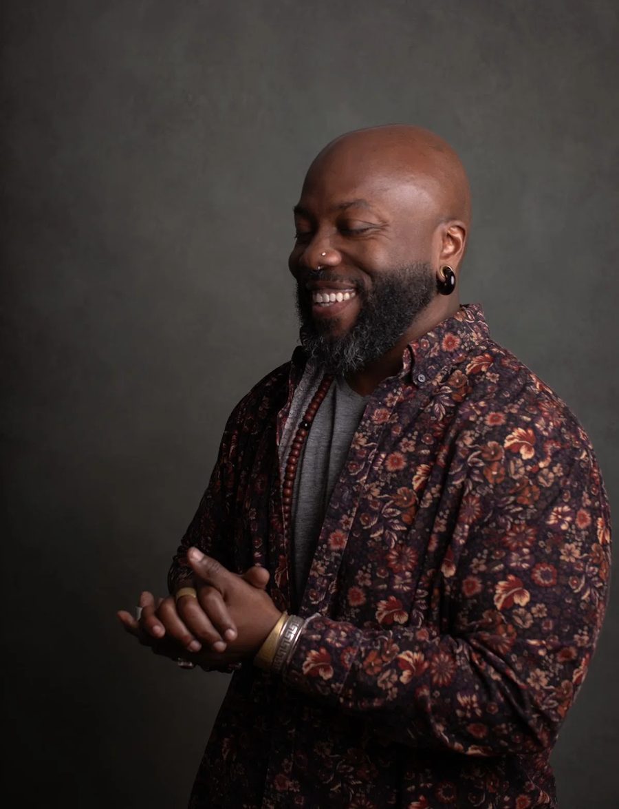 quincéy in a flowered shirt, turned aside and smiling wide, hands meeting.
