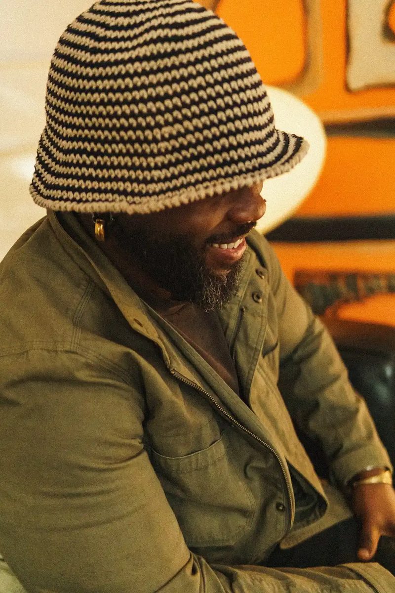 quincéy laughing beneath the brim of a striped crochet hat, orange glowing behind.