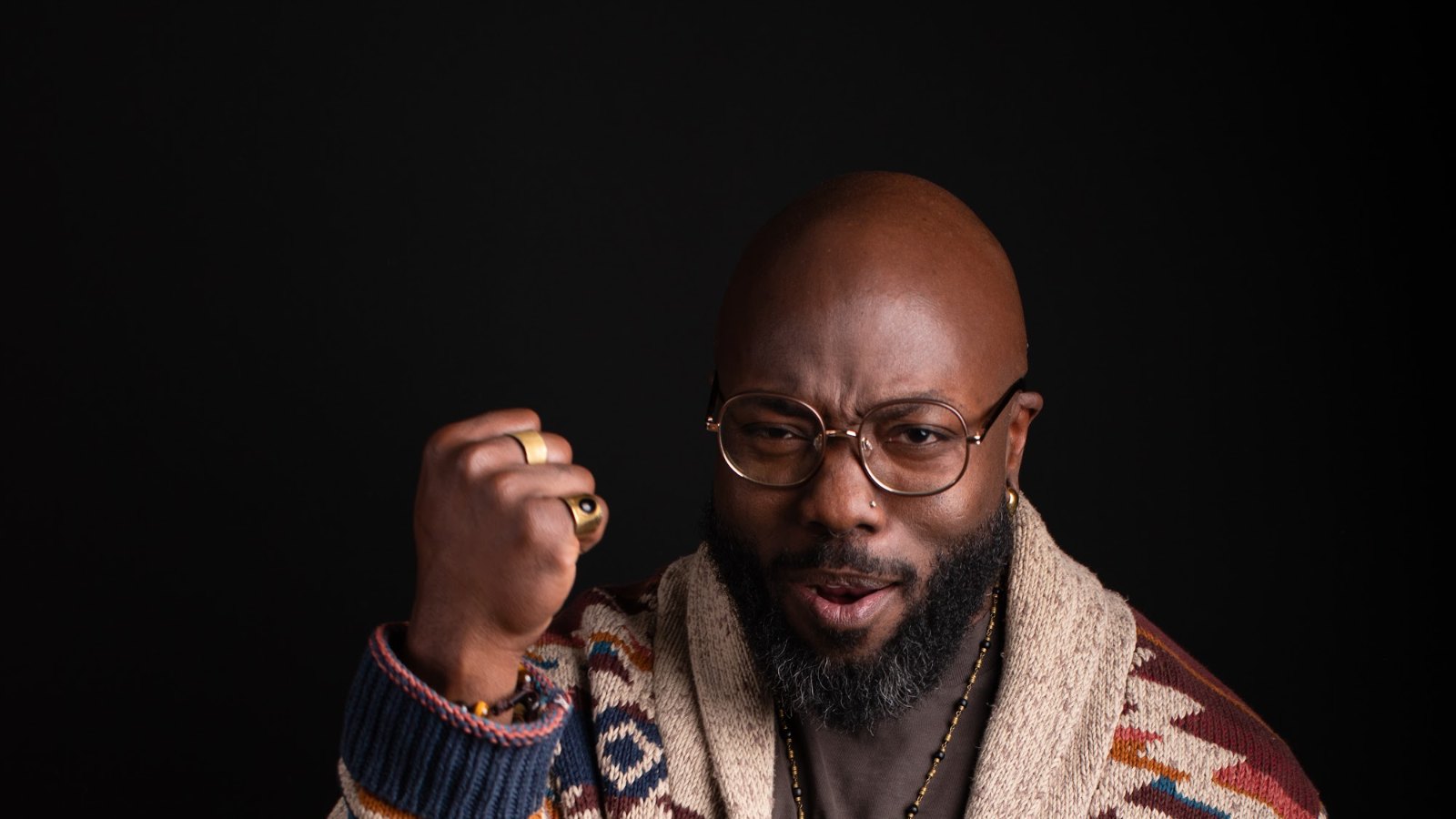 quincéy in a patterned cardigan and round glasses against a dark backdrop, right fist raised at the shoulder. photograph by owen m. hart.