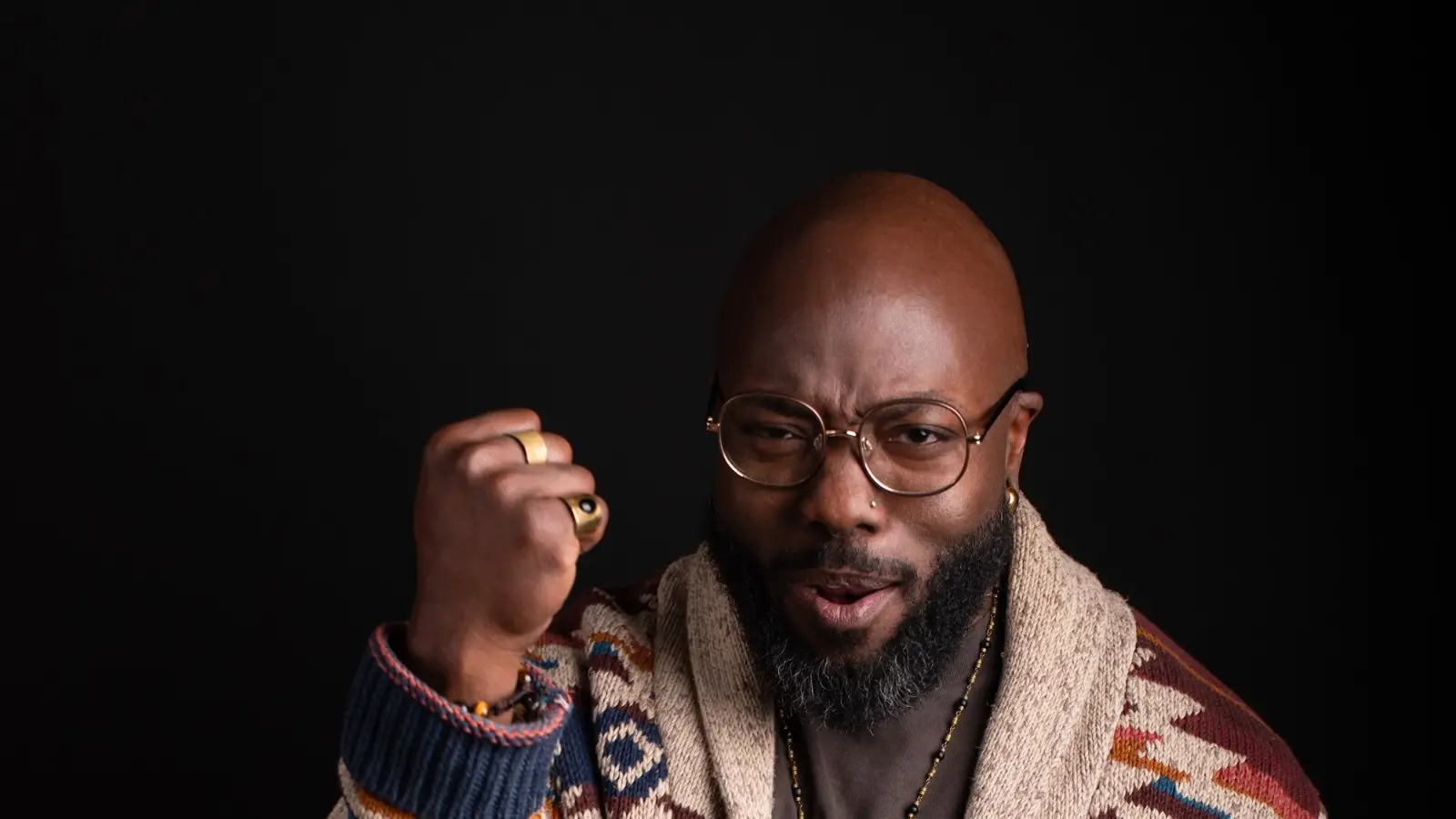 quincéy in a patterned cardigan and round glasses against a dark backdrop, right fist raised at the shoulder. photograph by owen m. hart.