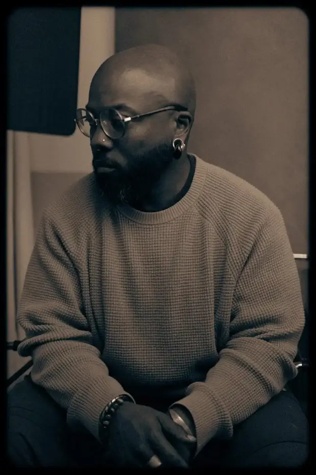 a muted film frame: quincéy in round glasses and a knit sweater, looking down and away, hands folded.