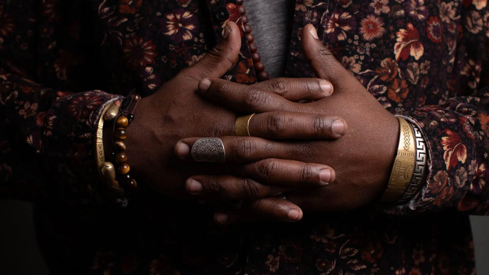 quincéy's hands clasped at the chest, rings and bracelets, against a floral corduroy shirt in low light.