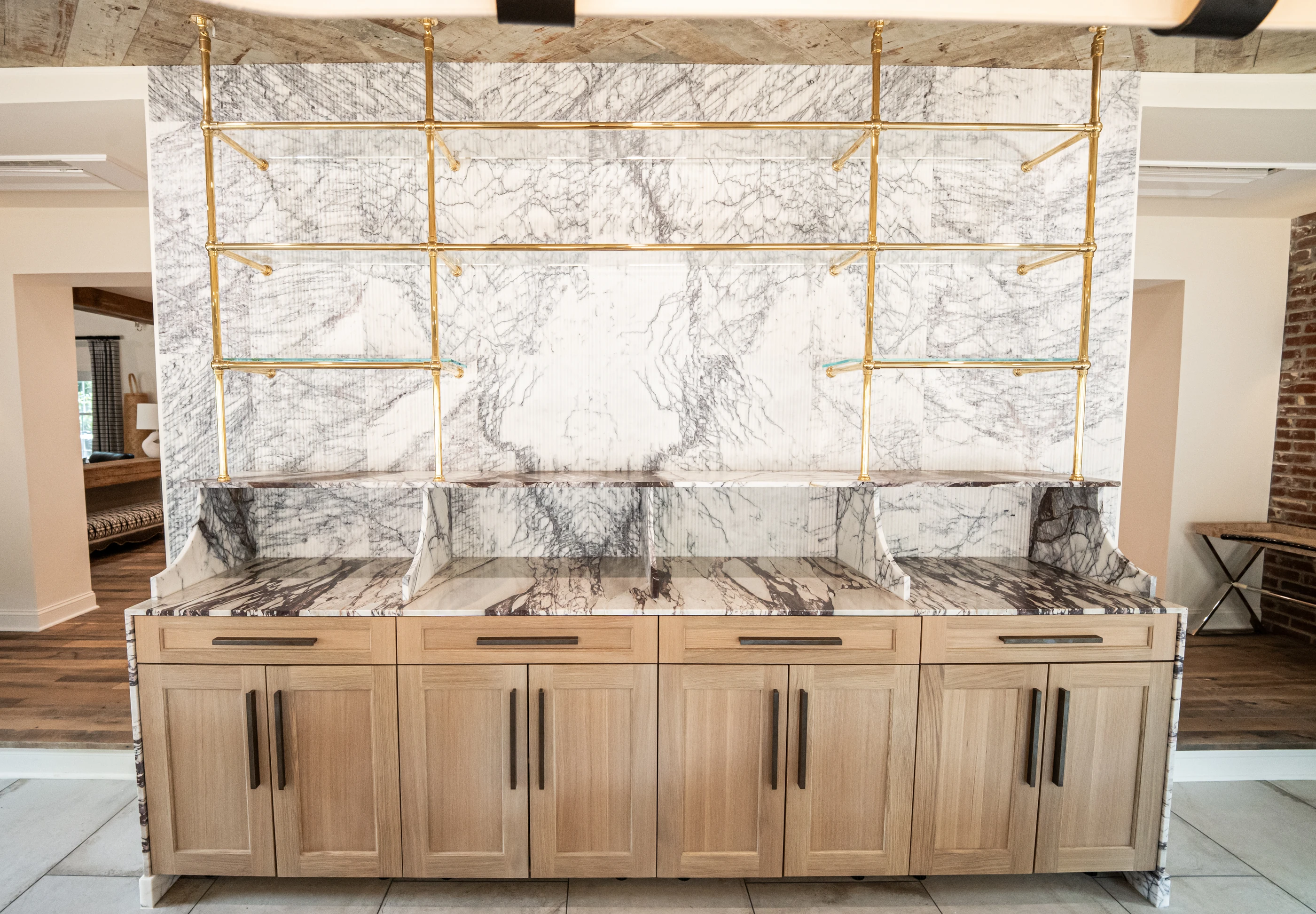 Hart stone and brass bar with veined marble, glass shelving and pale timber cabinetry.