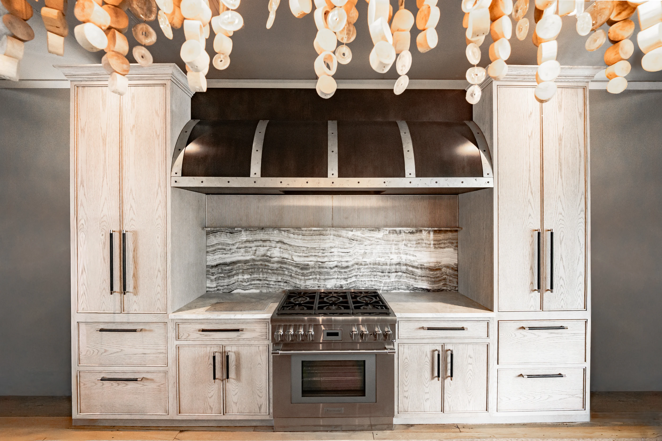 Pale kitchen cabinetry and sculptural metal range hood in the Cabinet & Stone Lab showroom.