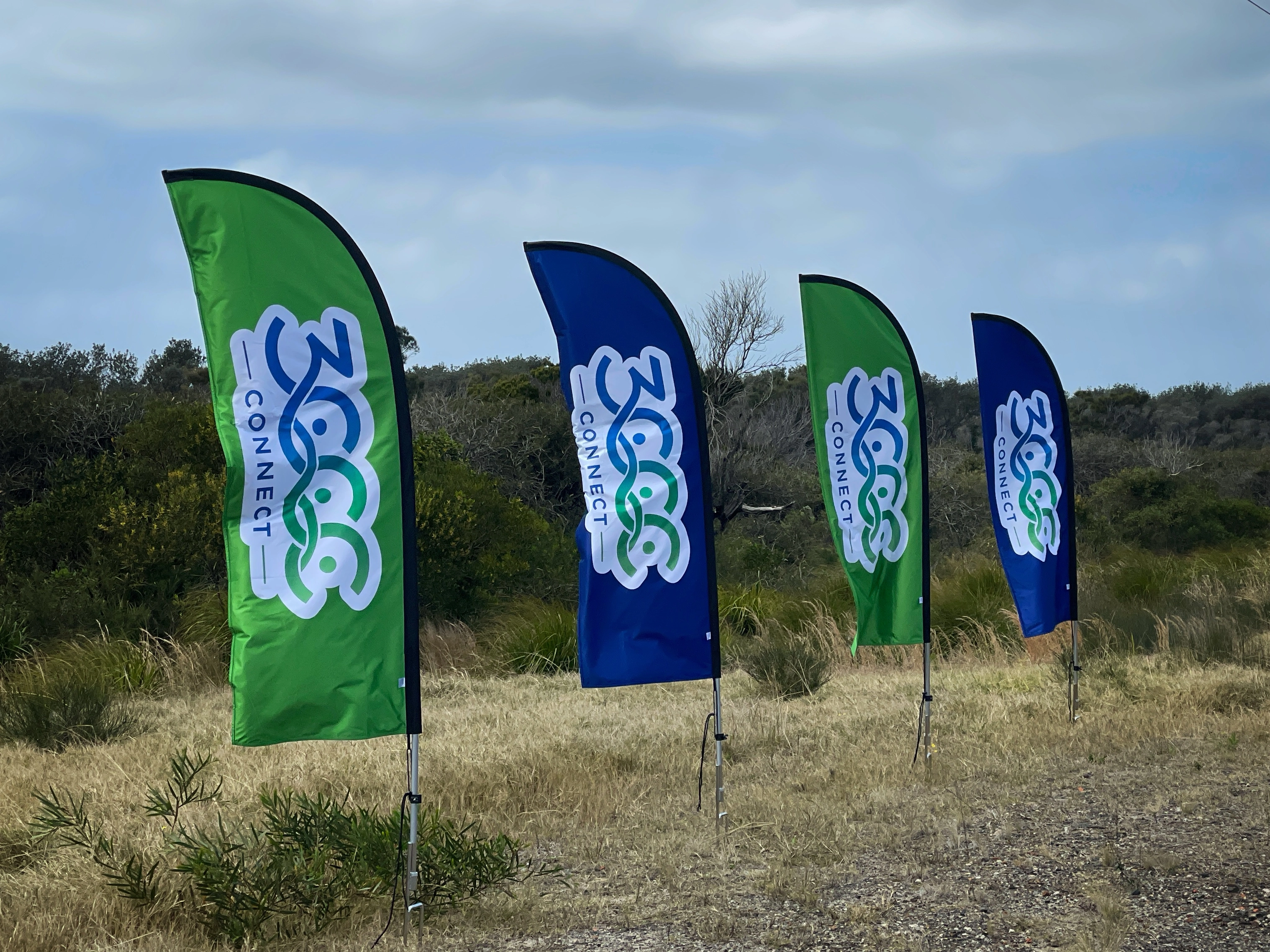 Image of 3C Connect banners along part of where the path is planned to be built. The location has short grass, a variety of bushes and some shrubs.