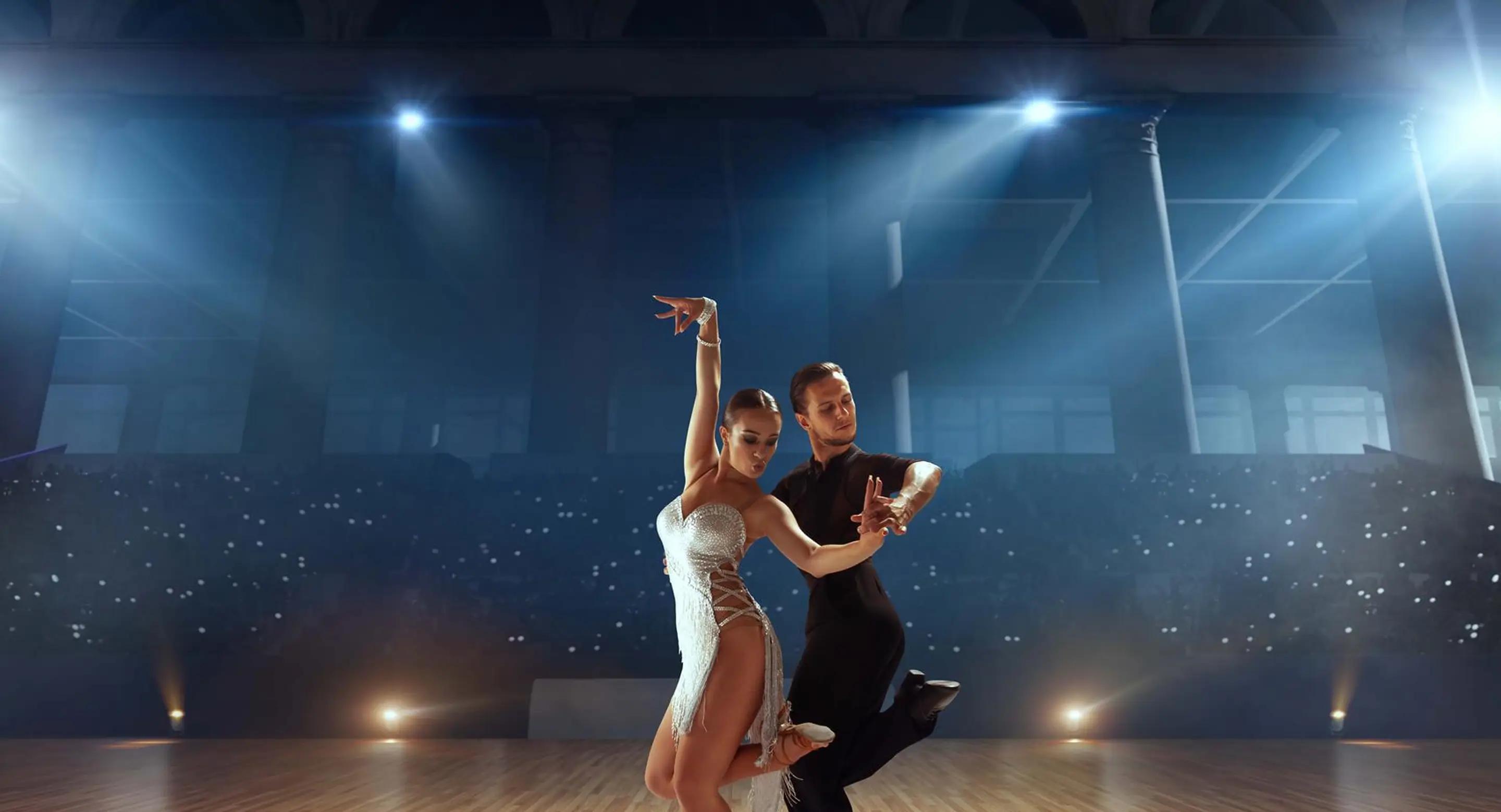 A professional dance couple performing a ballroom dance on a wooden floor under spotlights, with the female dancer in a white dress and the male dancer in black, against a dark audience background.