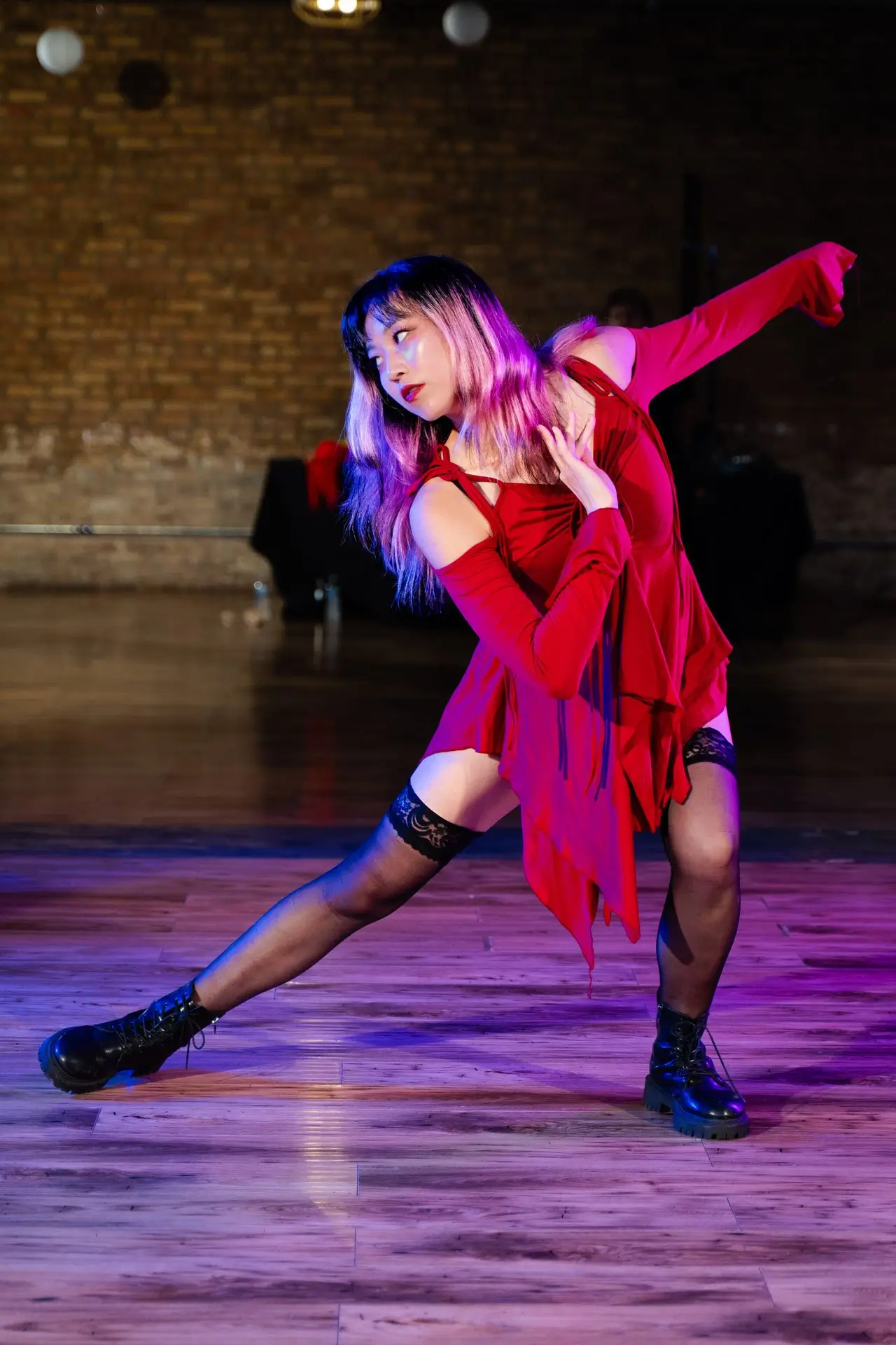 Woman with pink and blonde hair dressed in a red flowing outfit and black boots performing a dance pose on a wooden floor with a brick wall background.