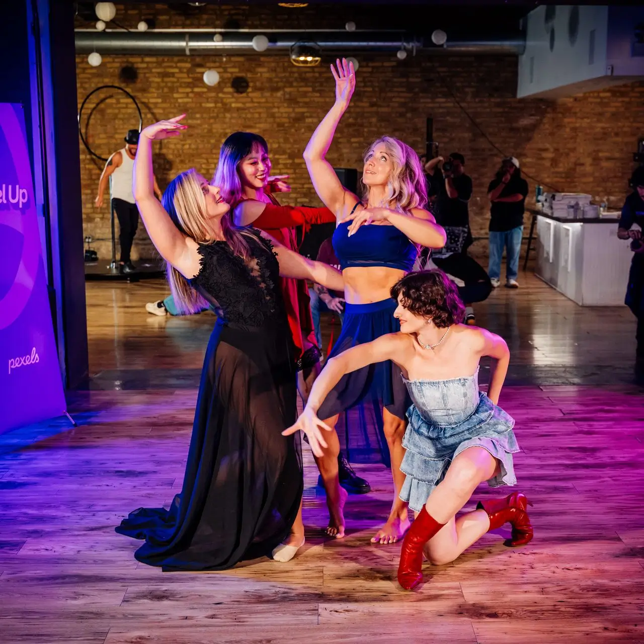 Four women in colorful outfits striking expressive dance poses on a wooden floor in an indoor studio with brick walls and purple lighting, while photographers and a man in the background observe.