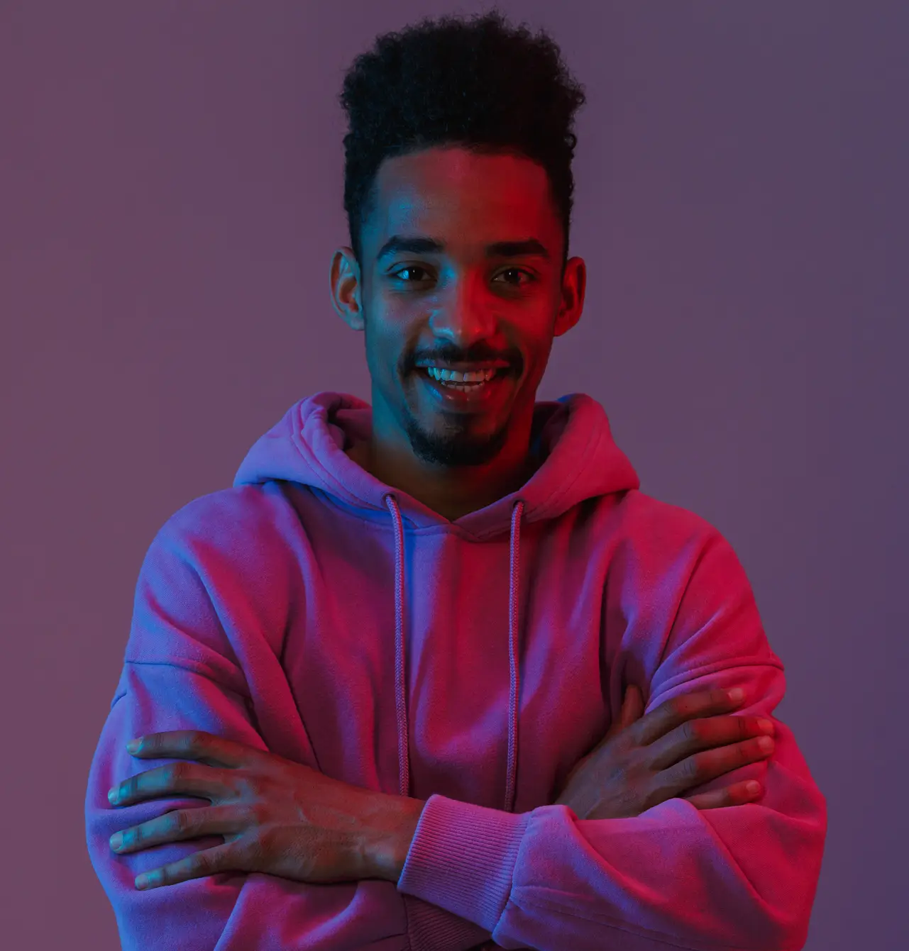 Man with curly dark hair and a goatee smiling with arms crossed, wearing a pink hoodie against a purple background with blue and pink lighting.