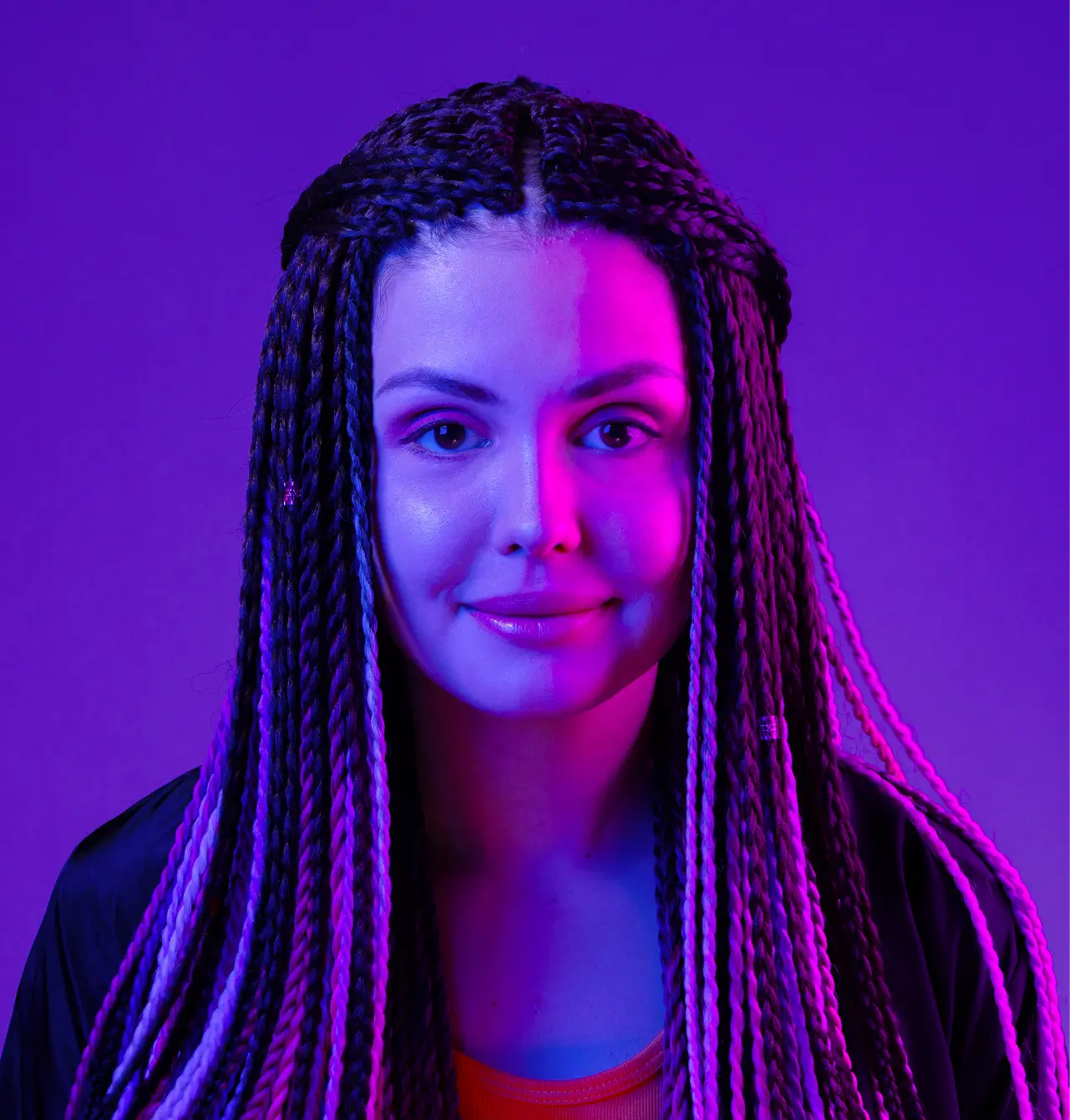 Portrait of a young woman with long braided hair, illuminated by vibrant purple and blue lighting, against a plain purple background.