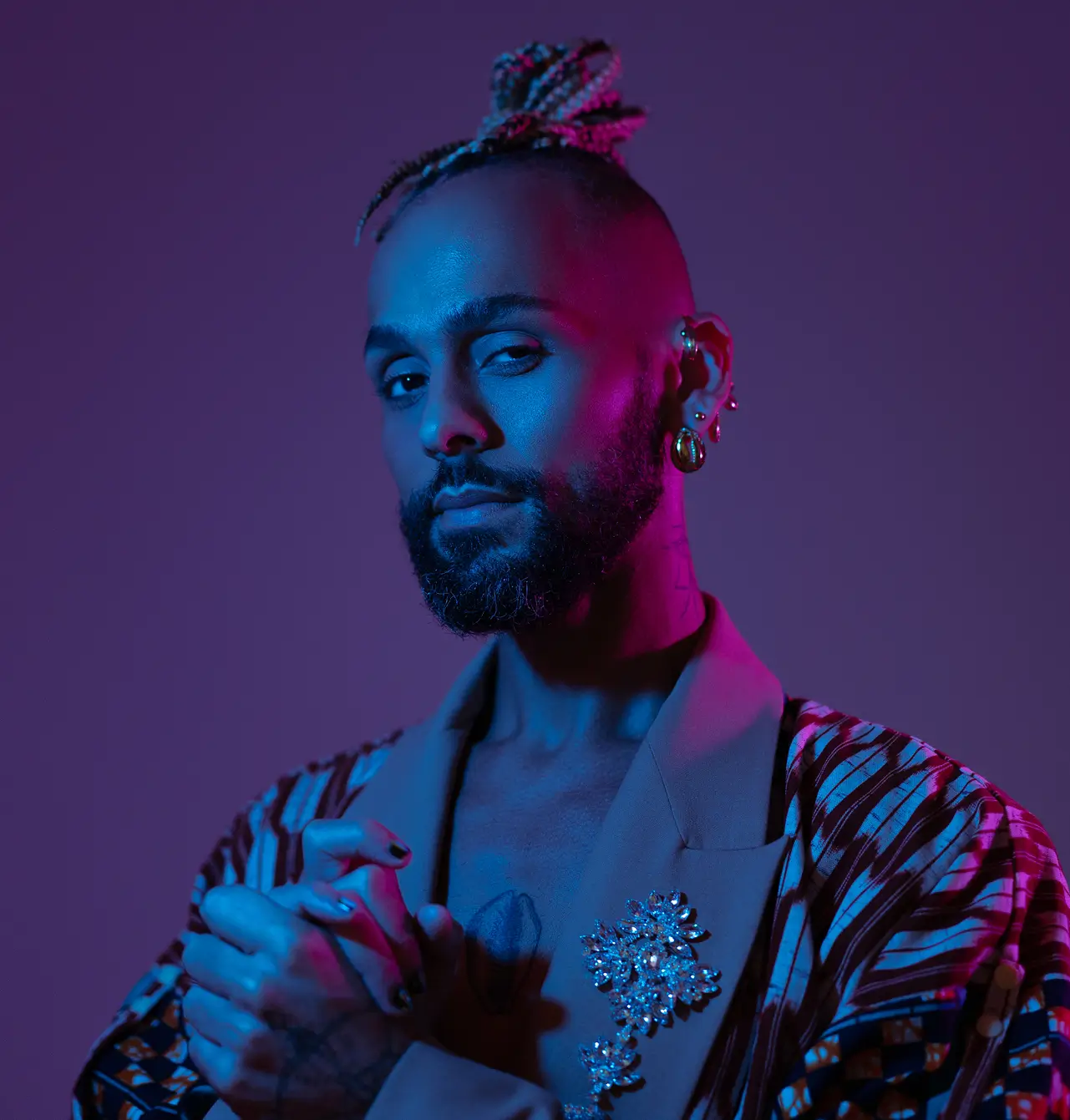 Portrait of a person with braided hair in a bun, a beard, and multiple earrings wearing a patterned jacket and blazer with jeweled brooches, illuminated by blue and pink lighting against a purple background.