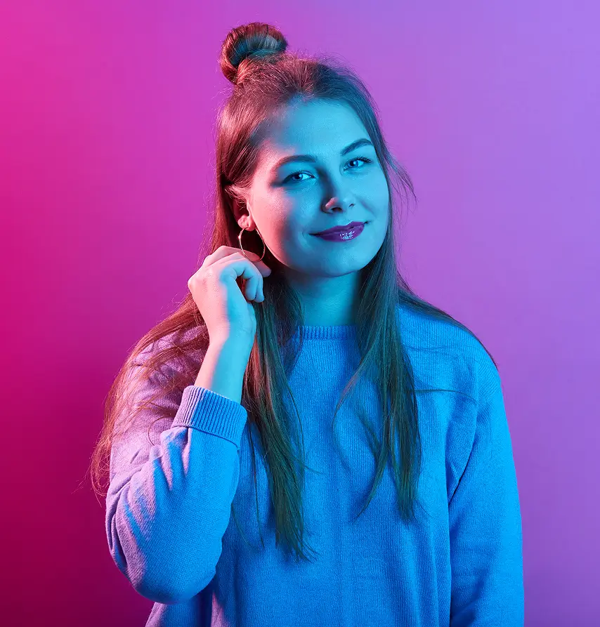 Young woman with long brown hair partially styled in a top bun, wearing a light blue sweater and hoop earrings, smiling slightly against a pink and purple gradient background with blue lighting on her face and upper body.