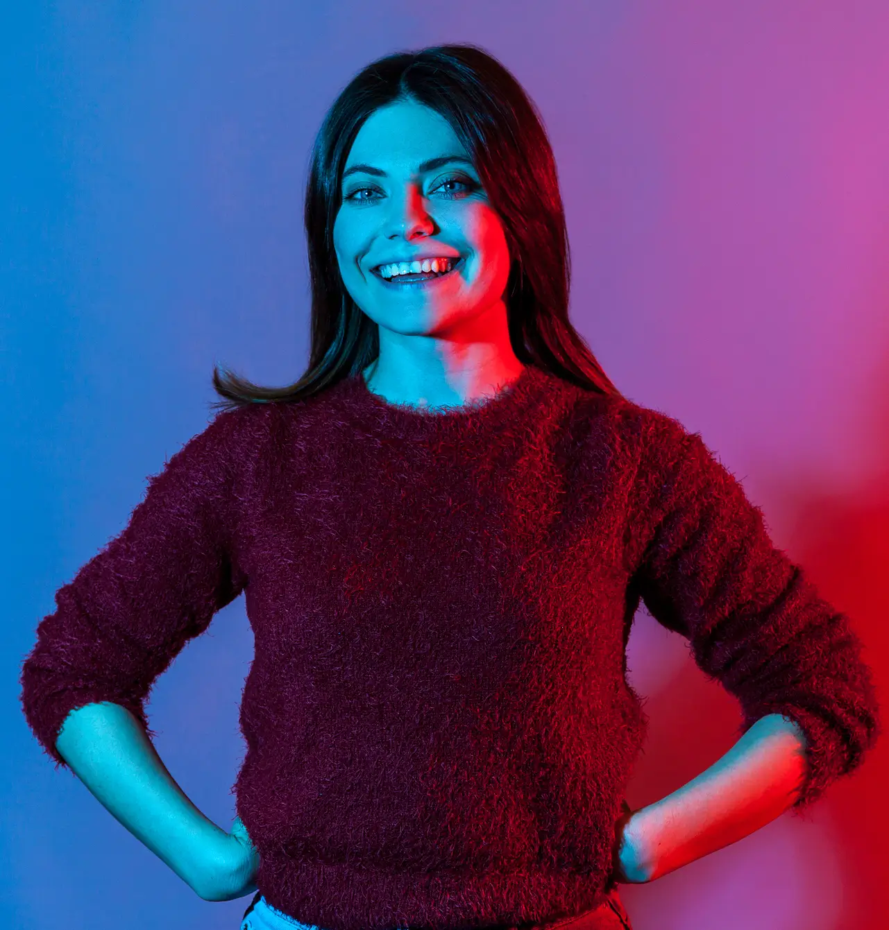 Smiling woman with dark hair wearing a fuzzy burgundy sweater, standing with hands on hips against a background lit with blue and red lighting.