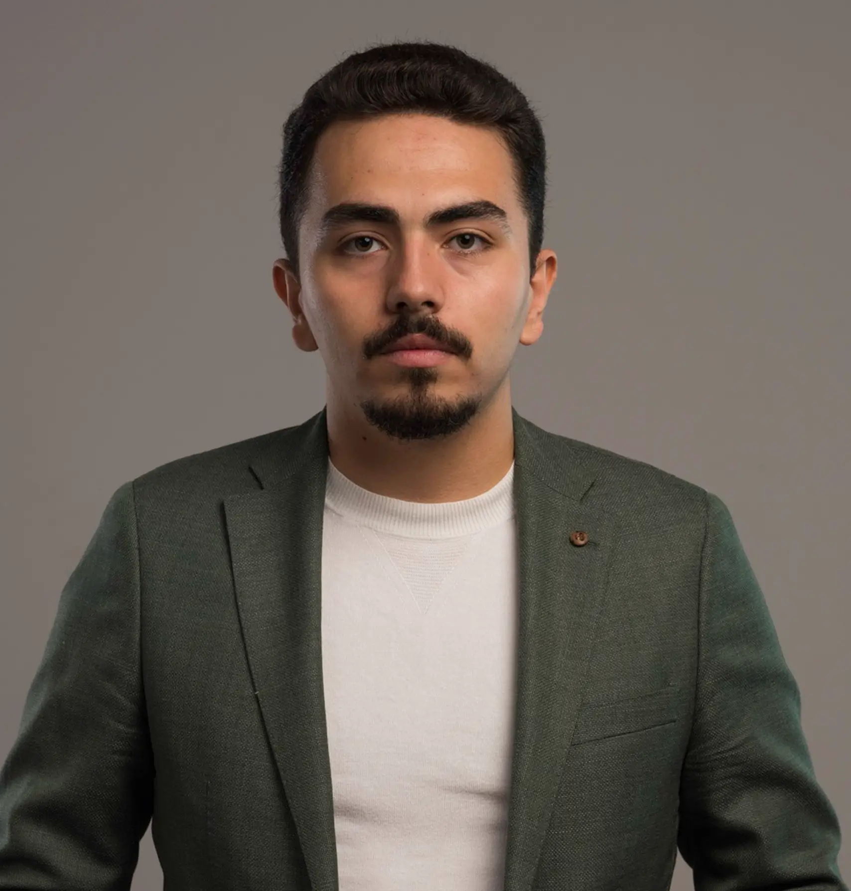 Portrait of a serious young man with dark hair and a short beard wearing a green blazer and white shirt against a plain gray background.