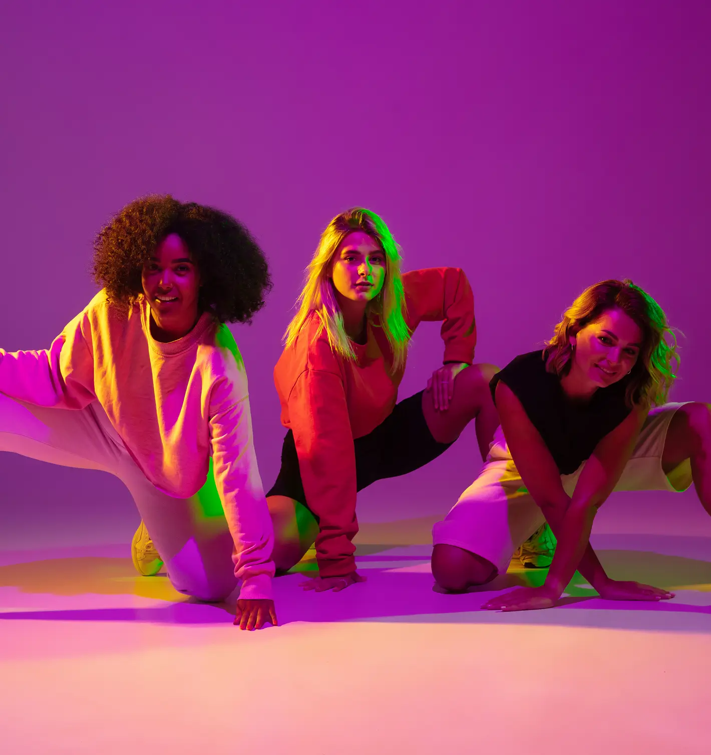 Three young women posing on the ground in a dance or athletic stance under colorful pink, purple, yellow, and green studio lighting, against a plain purple background.