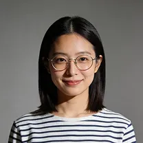 Portrait of a young Asian woman with straight black hair wearing round glasses and a white shirt with black horizontal stripes, smiling against a plain gray background.