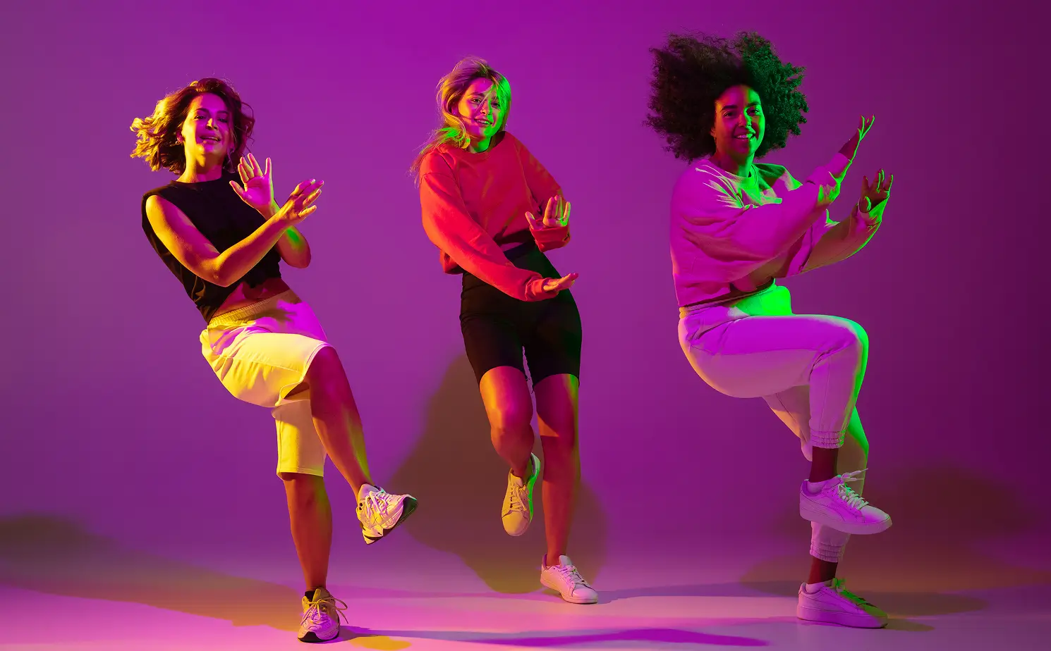 Three women dancing energetically against a purple background with colorful lighting, each lifting one leg and smiling.