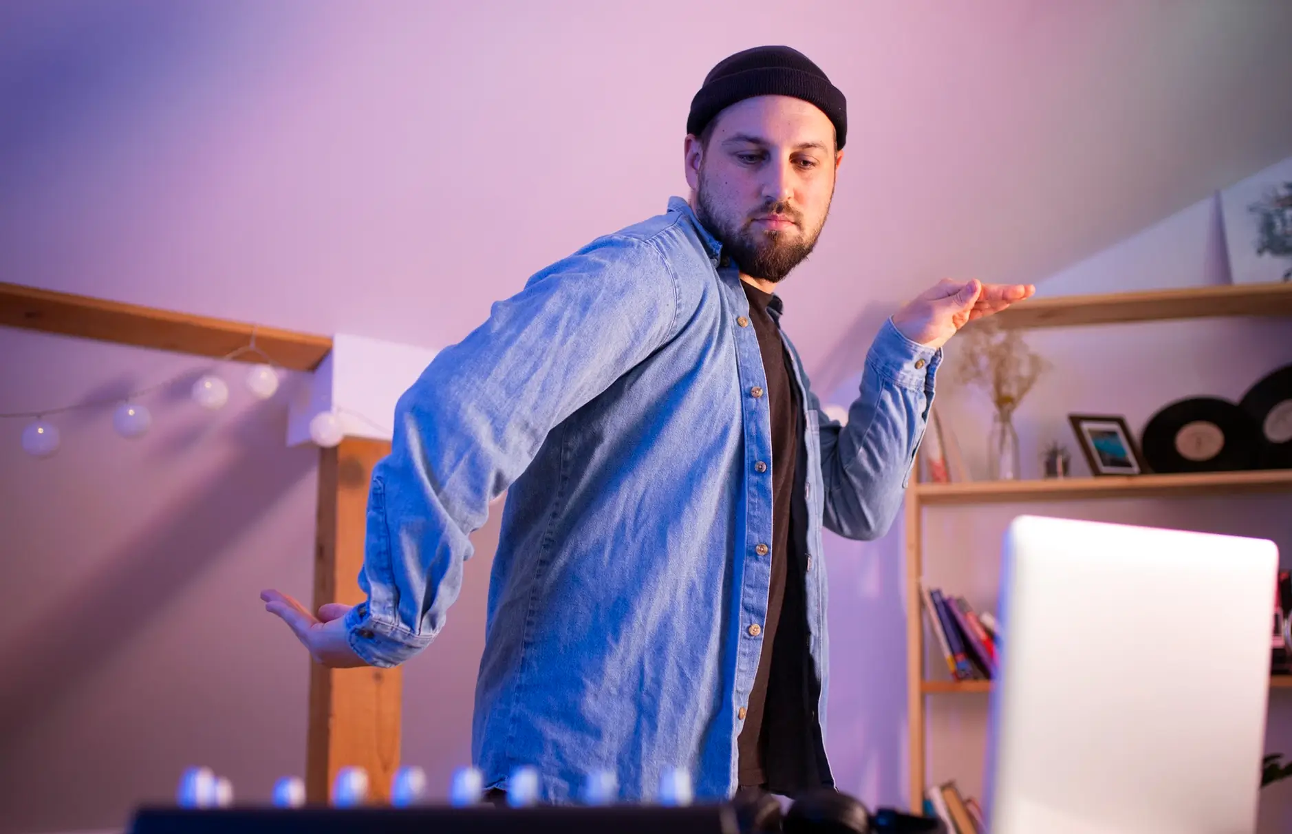 A man wearing a black beanie and denim shirt stands in a room with shelves in the background, appearing to DJ or mix music using equipment and a laptop in front of him.