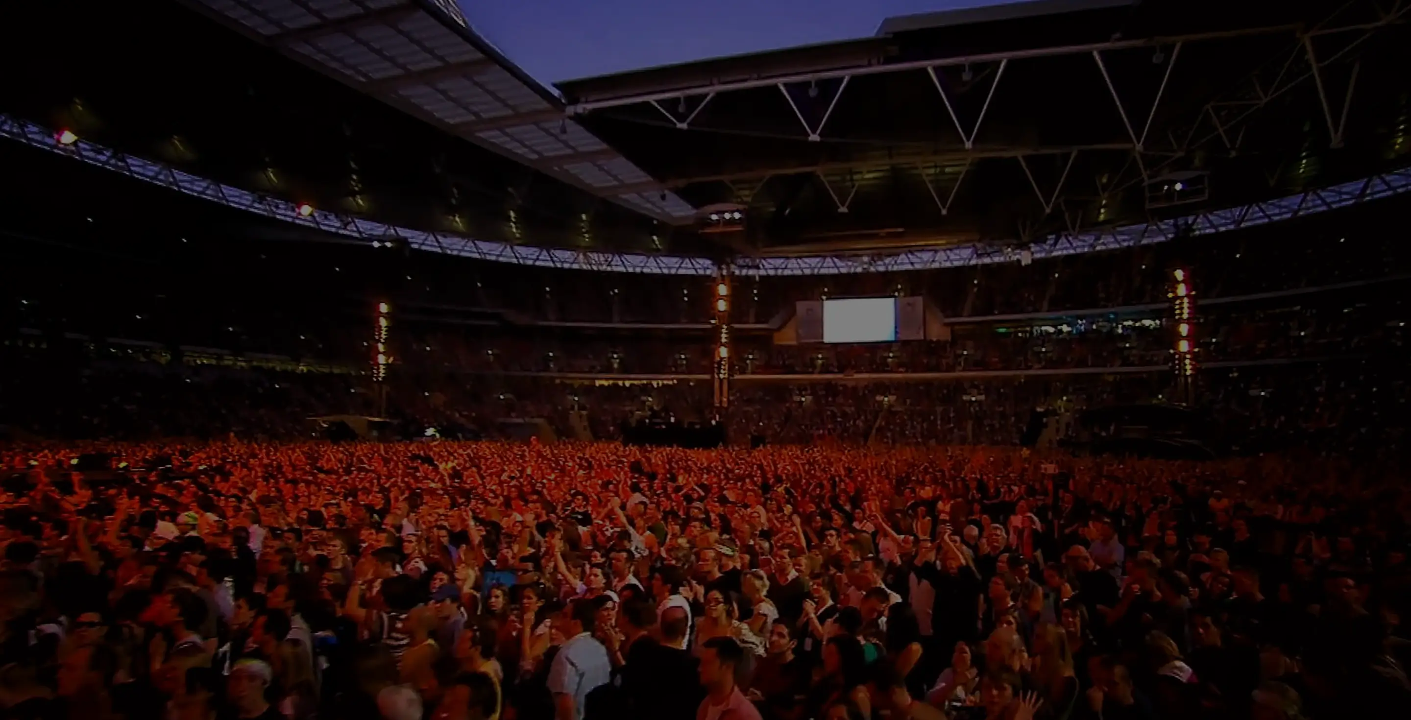 Large crowd of people illuminated by orange stage lights inside a stadium with a partially open roof during an event.