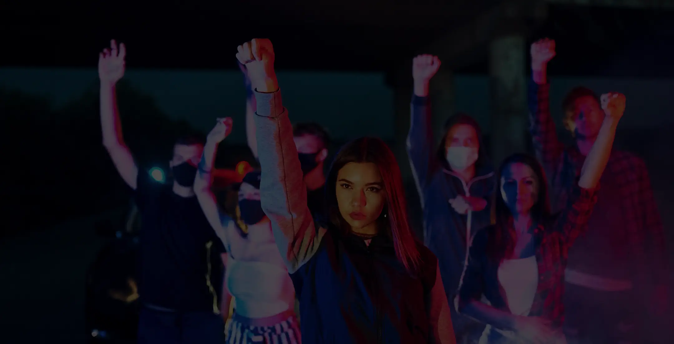 Group of young people standing outdoors at night with raised fists, some wearing face masks, expressing solidarity or protest.