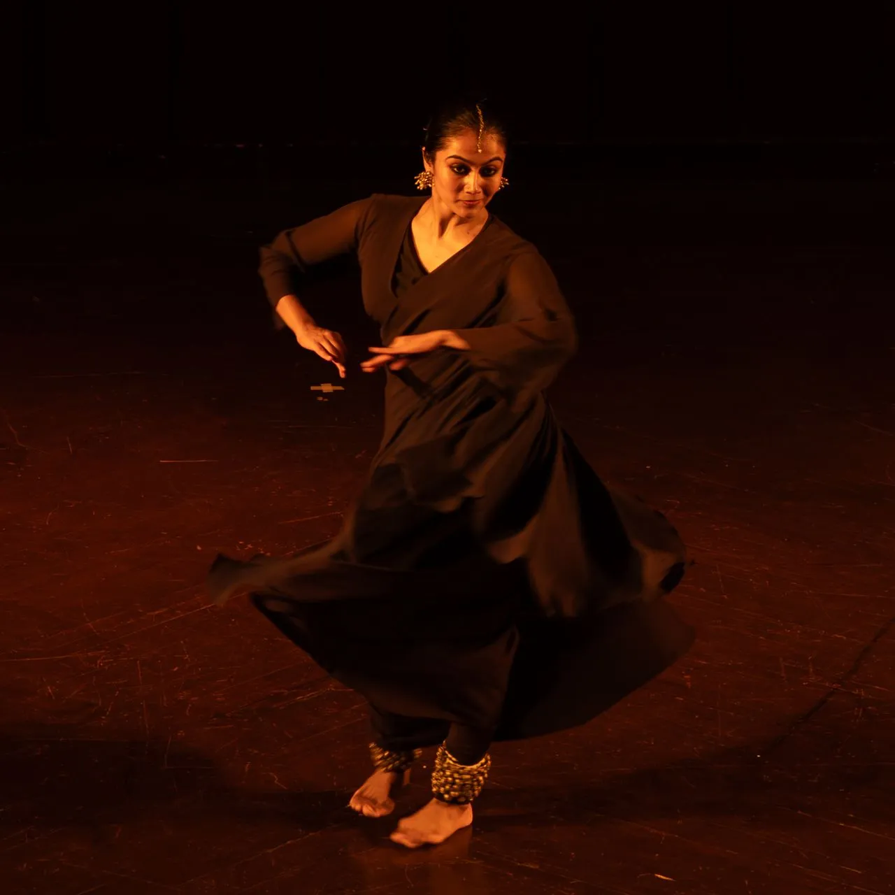 A woman in traditional Indian dance attire with ankle bells performs a classical dance on a dimly lit stage, her flowing dress swirling as she moves.