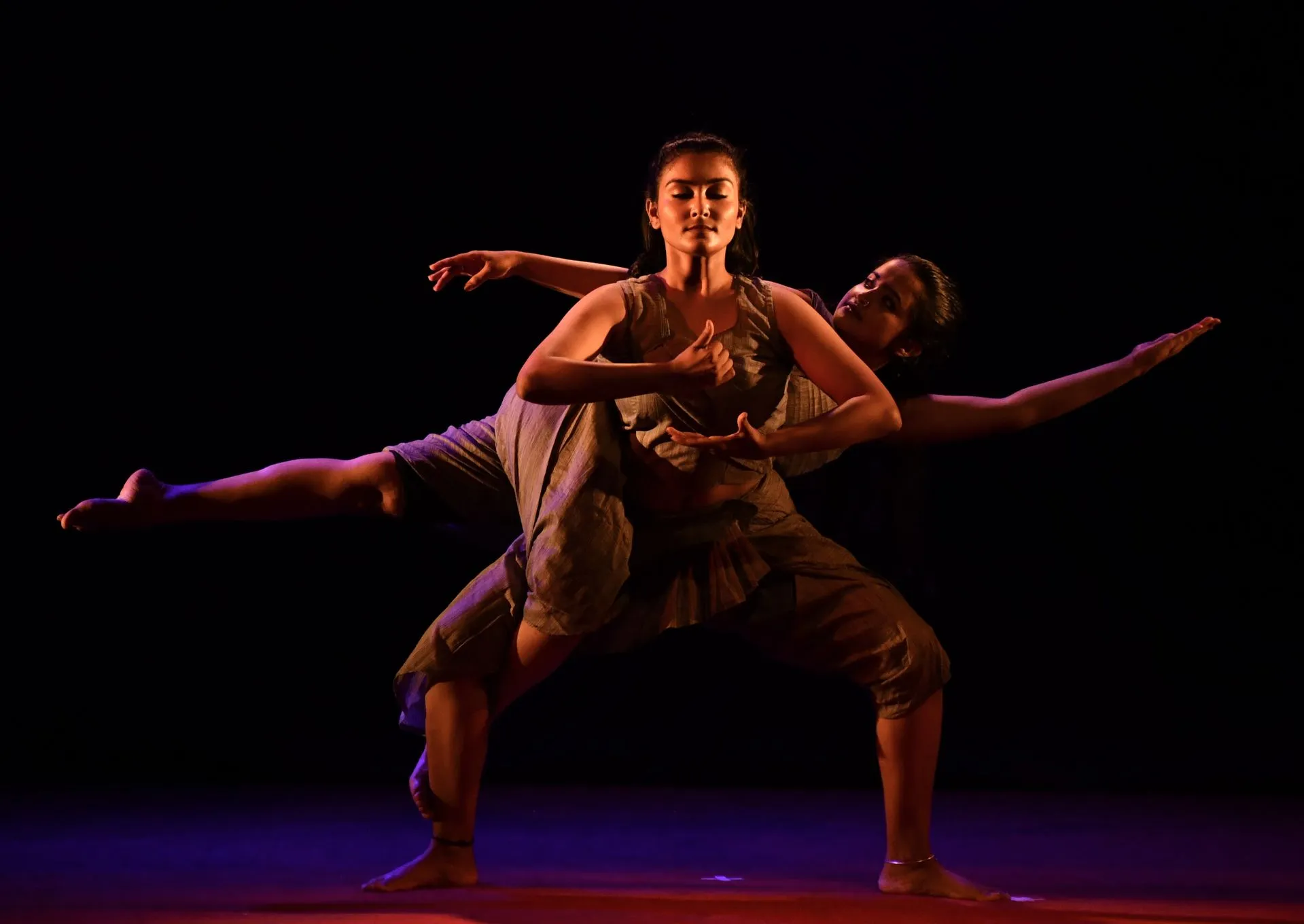 Two female dancers performing a contemporary dance pose on stage with dramatic lighting against a black background, wearing loose, earthy-toned outfits.