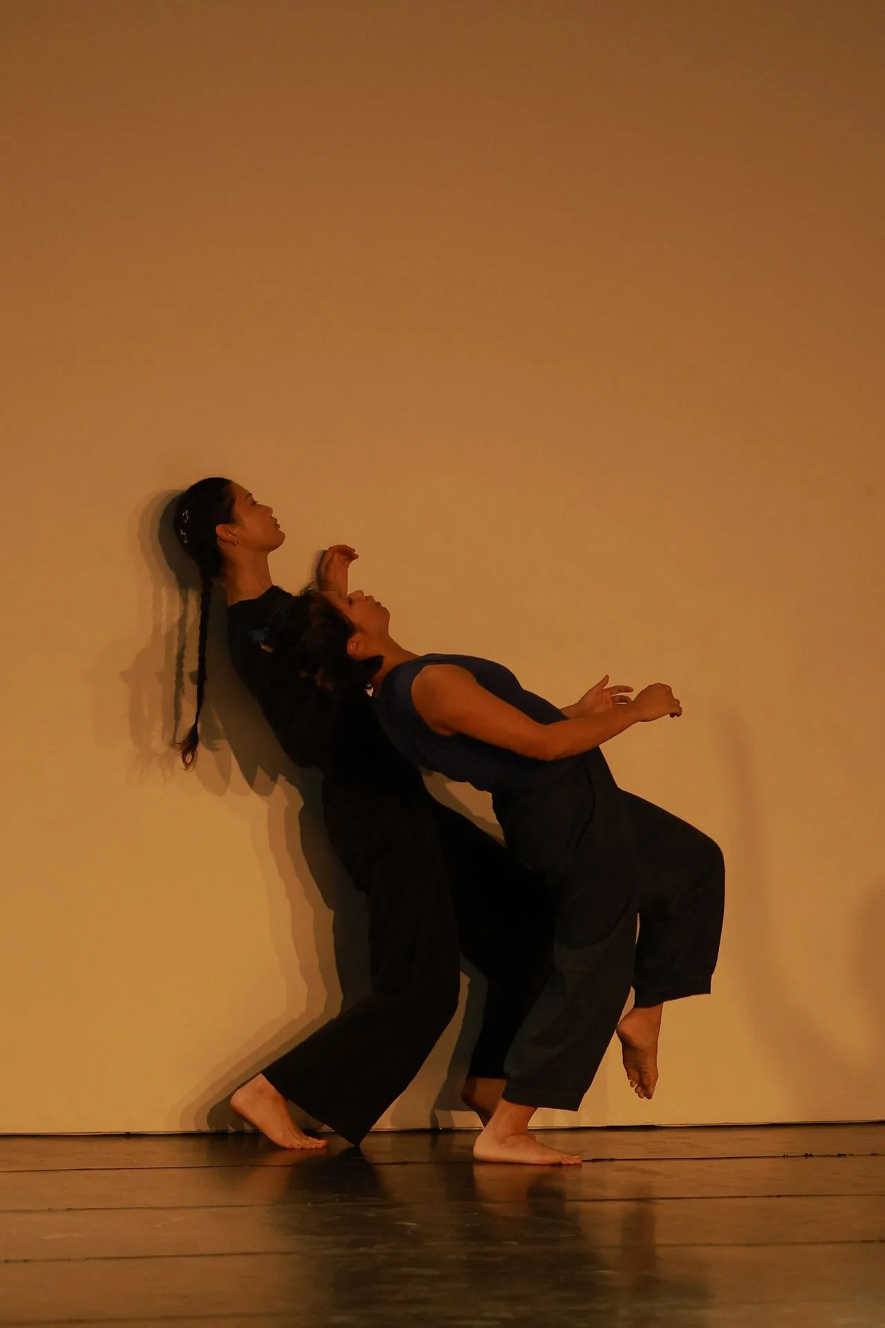 Two barefoot dancers in loose dark clothing perform a contemporary dance against a plain beige background, leaning backward with coordinated poses and shadows on the wall behind them.