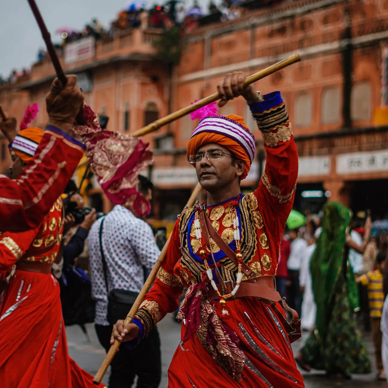 Man dressed in vibrant traditional Indian attire with red and gold embroidery, wearing an orange turban with purple and white stripes, holding a decorated wooden stick during a cultural street celebration.