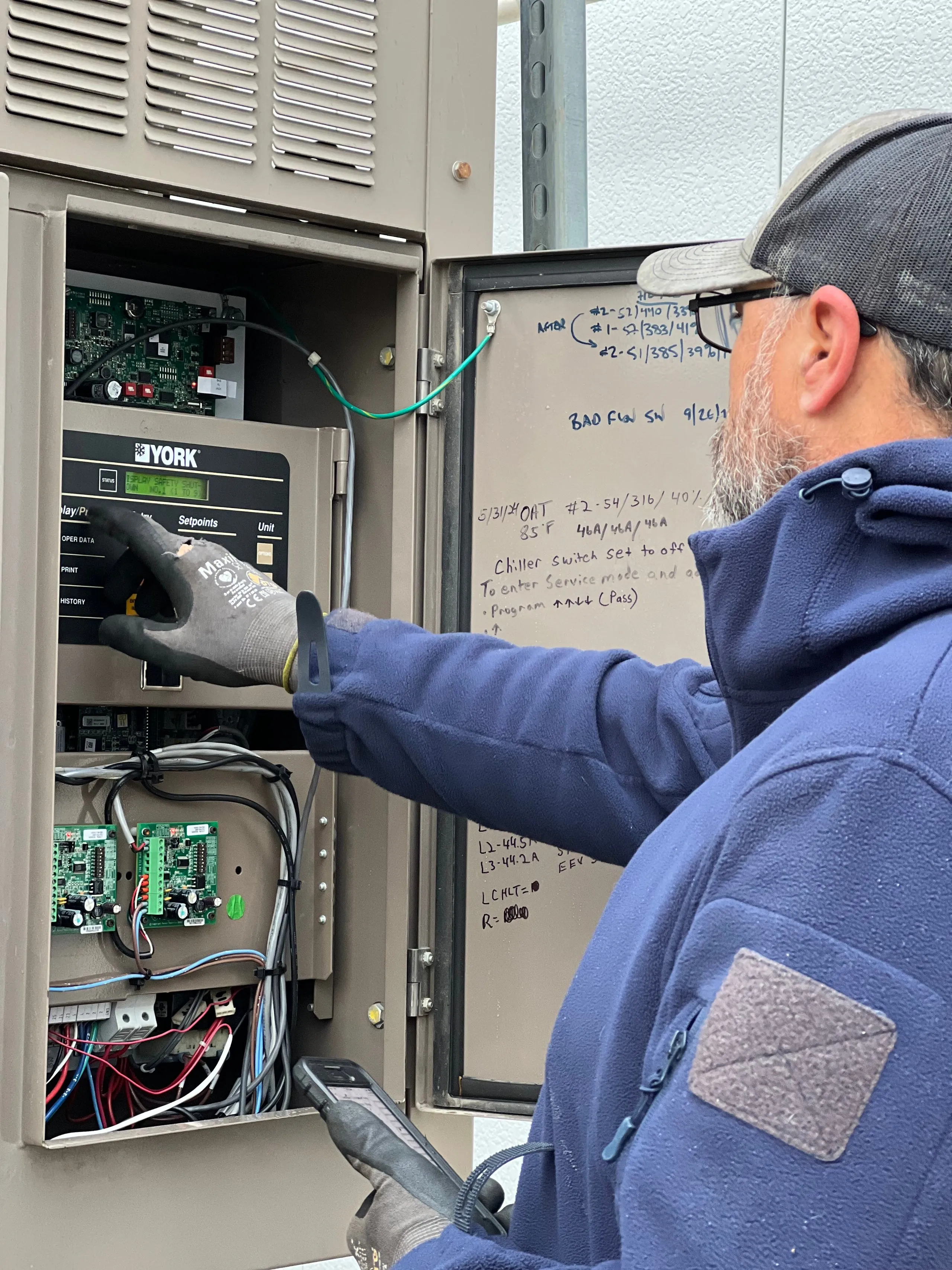 Technician wearing a navy blue jacket and gray gloves operates a York HVAC control panel inside an open metal electrical box with circuit boards and wires, while holding a handheld device and examining handwritten notes on the panel's door.