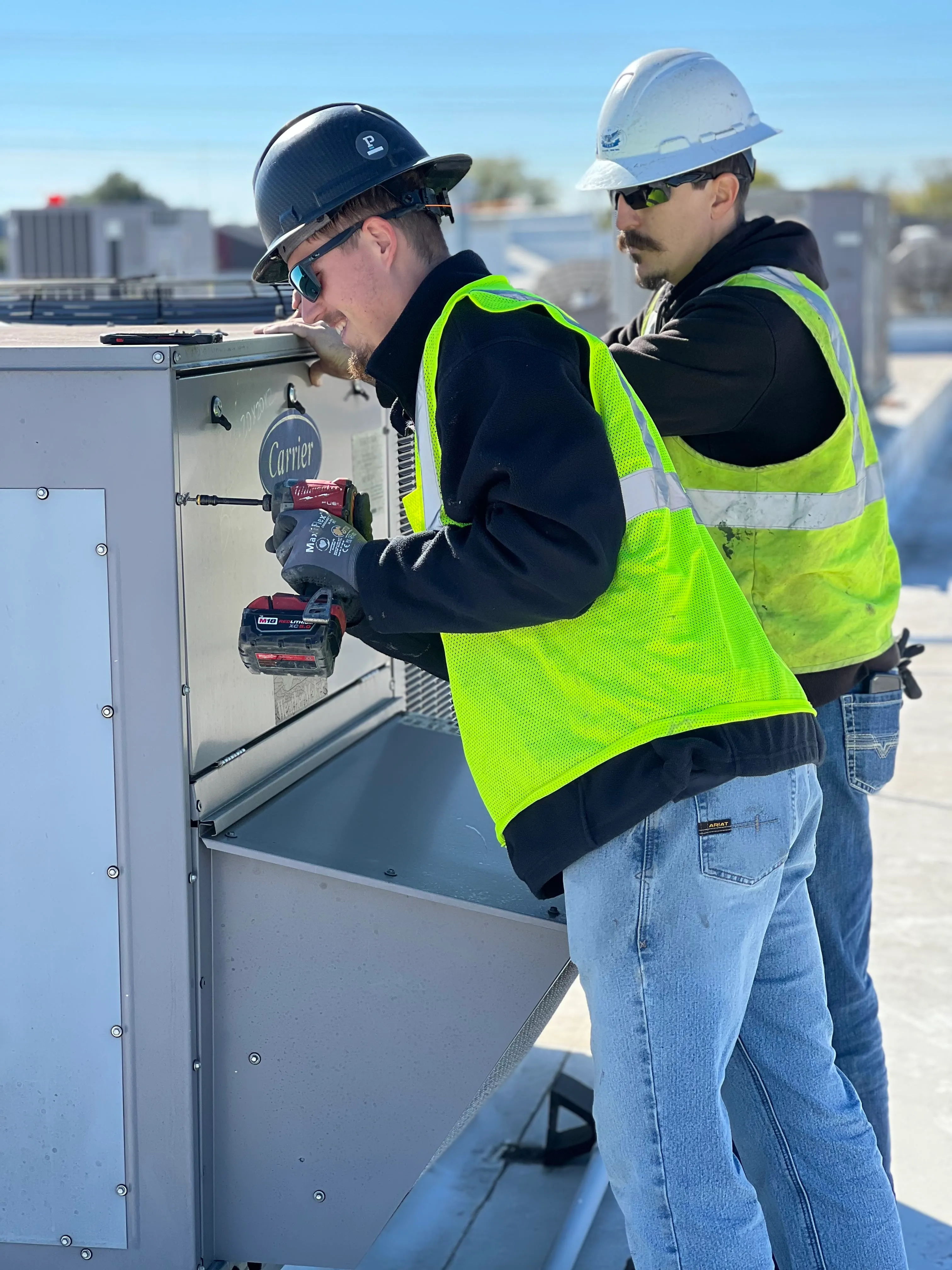 Two construction workers wearing hard hats, safety glasses, and high-visibility vests working on a rooftop HVAC unit, one using a power drill on the unit's panel.