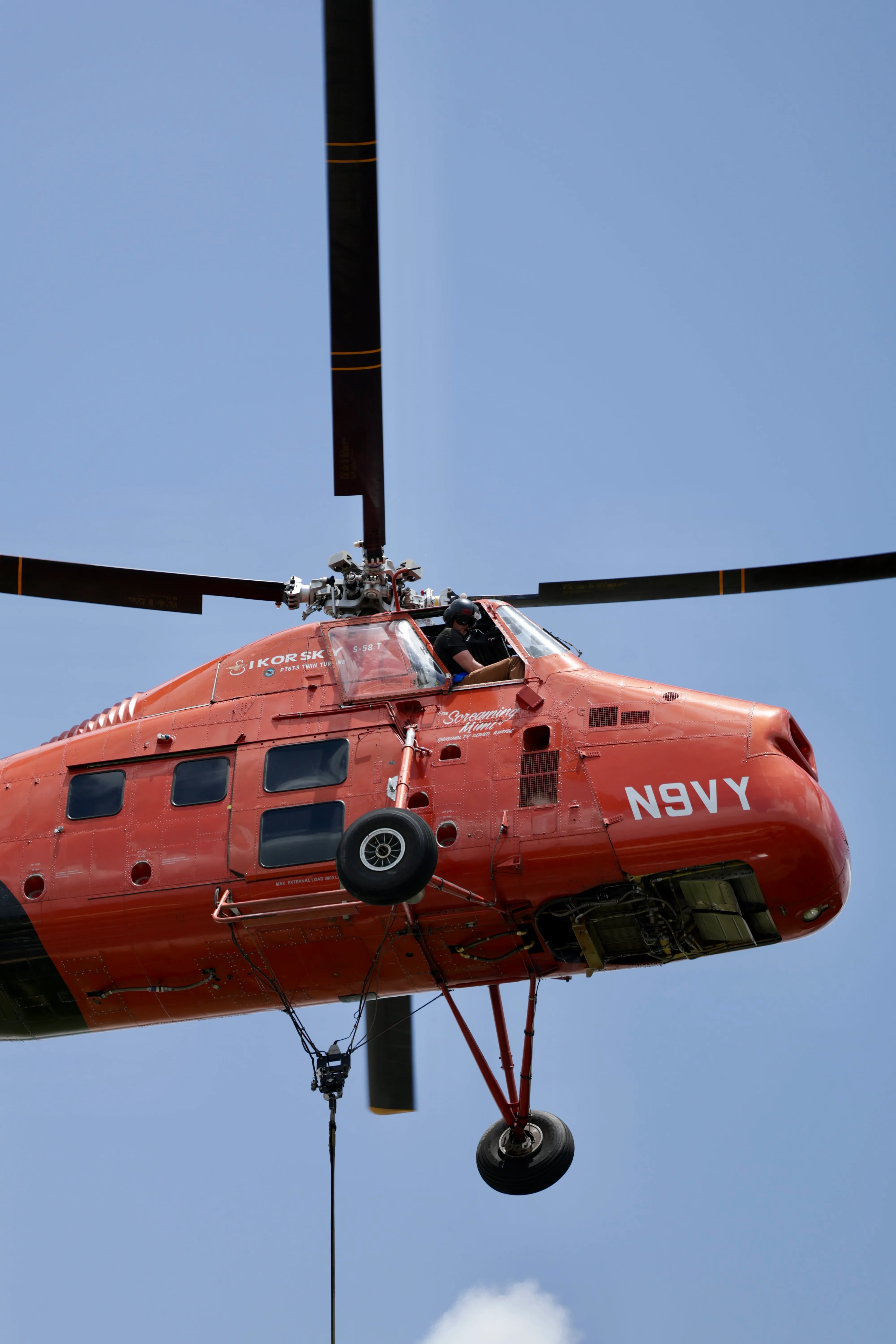 Red Sikorsky S-58T helicopter with a pilot in the cockpit, hovering against a clear blue sky, equipped with landing gear and a cable hanging from its undercarriage.