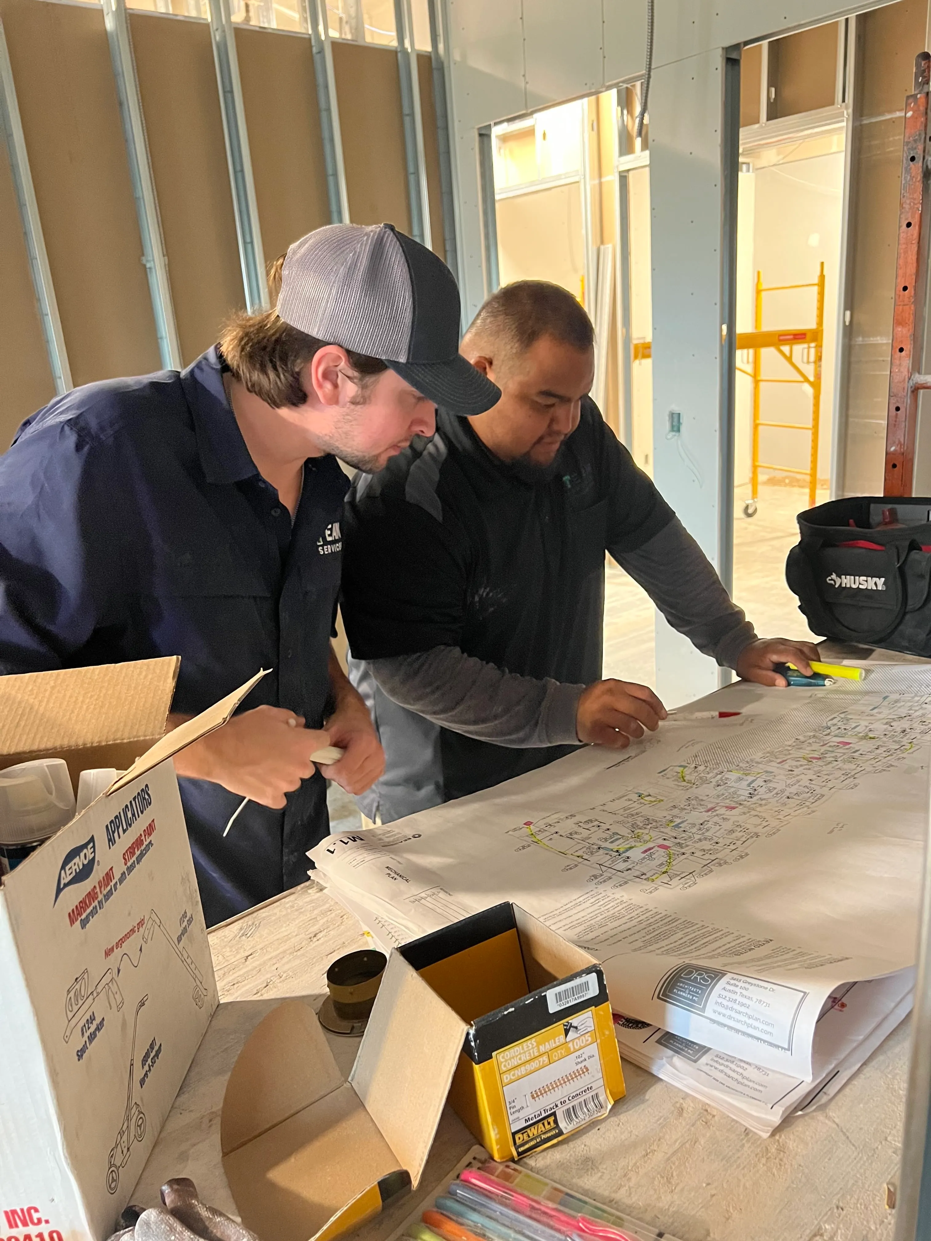 Two men examining a large blueprint spread out on a table at a construction site, with boxes of tools and colored markers nearby.
