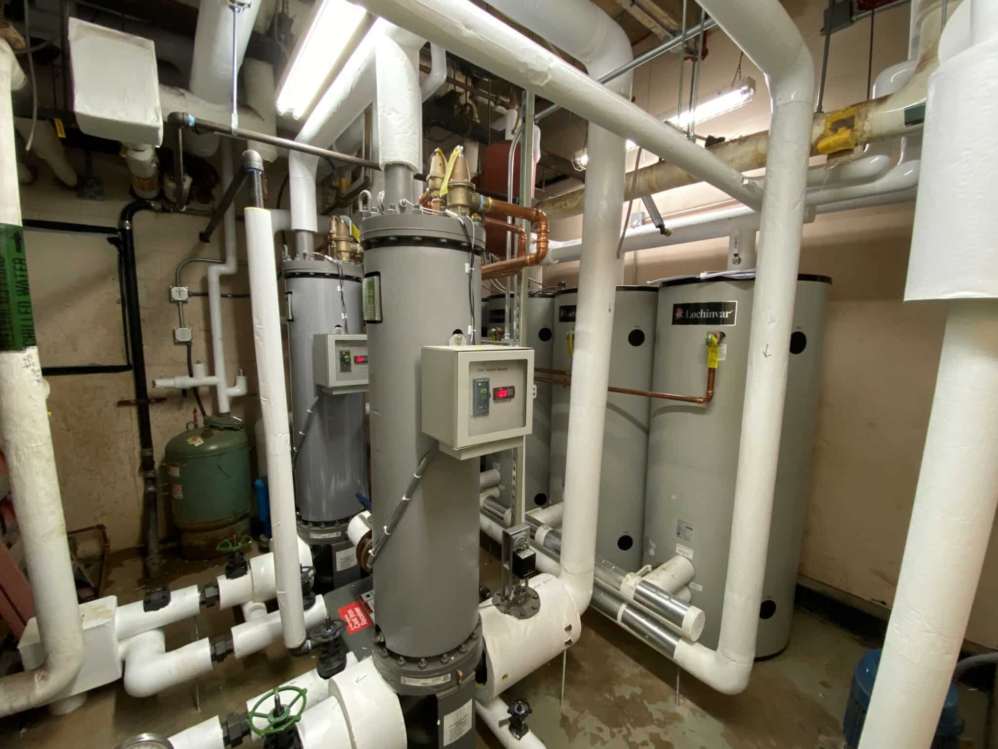 Industrial boiler room with multiple large cylindrical tanks and insulated pipes interconnected, control panels attached to tanks, floor wet with some puddles, beige walls under fluorescent lighting.