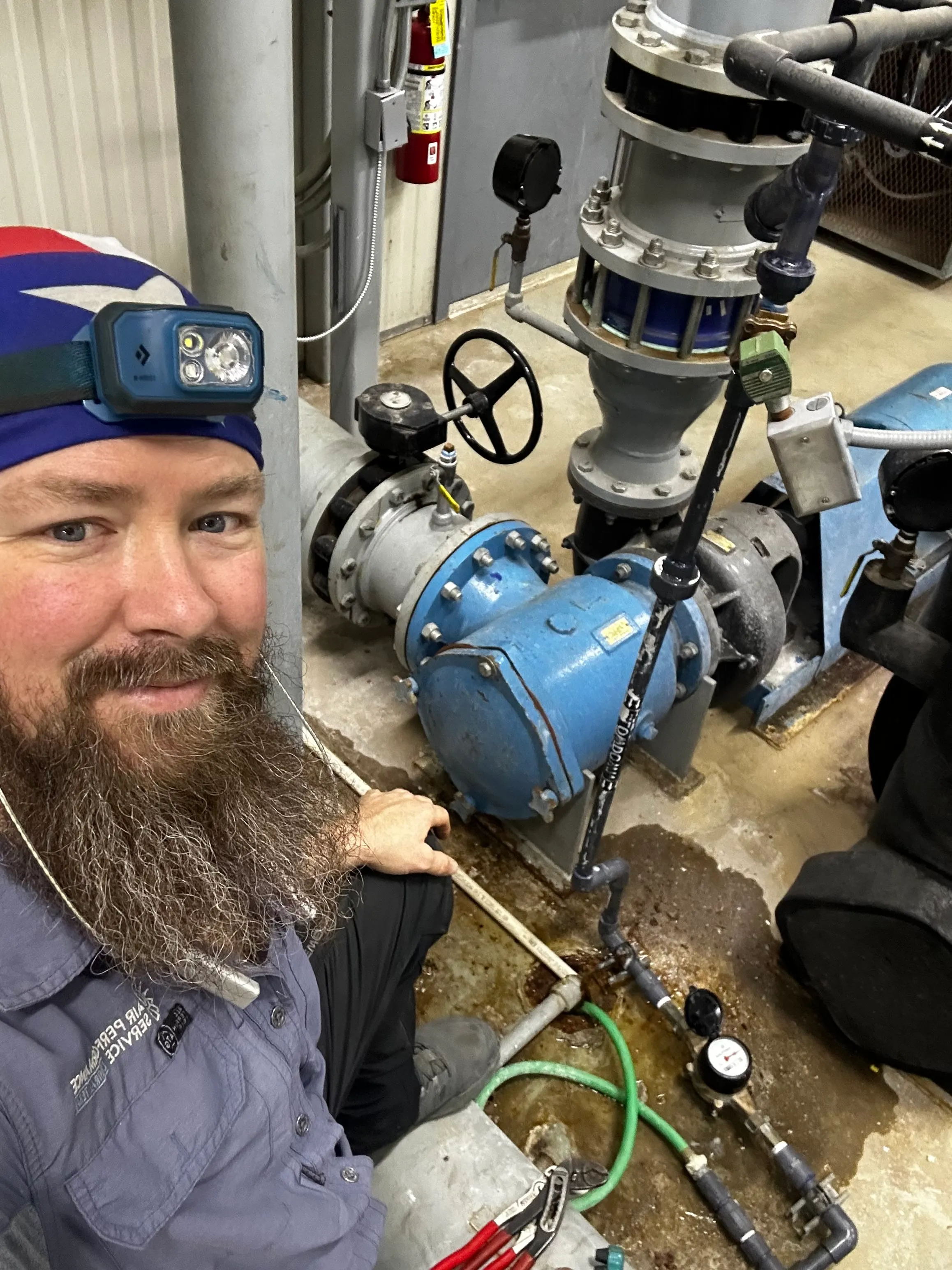 Bearded man wearing a blue headlamp and blue work shirt with a patch reading 'AIR PERFORMANCE SERVICE' sits next to large industrial blue pump machinery and pipes on a concrete floor, holding a tool with another tool on the floor nearby.