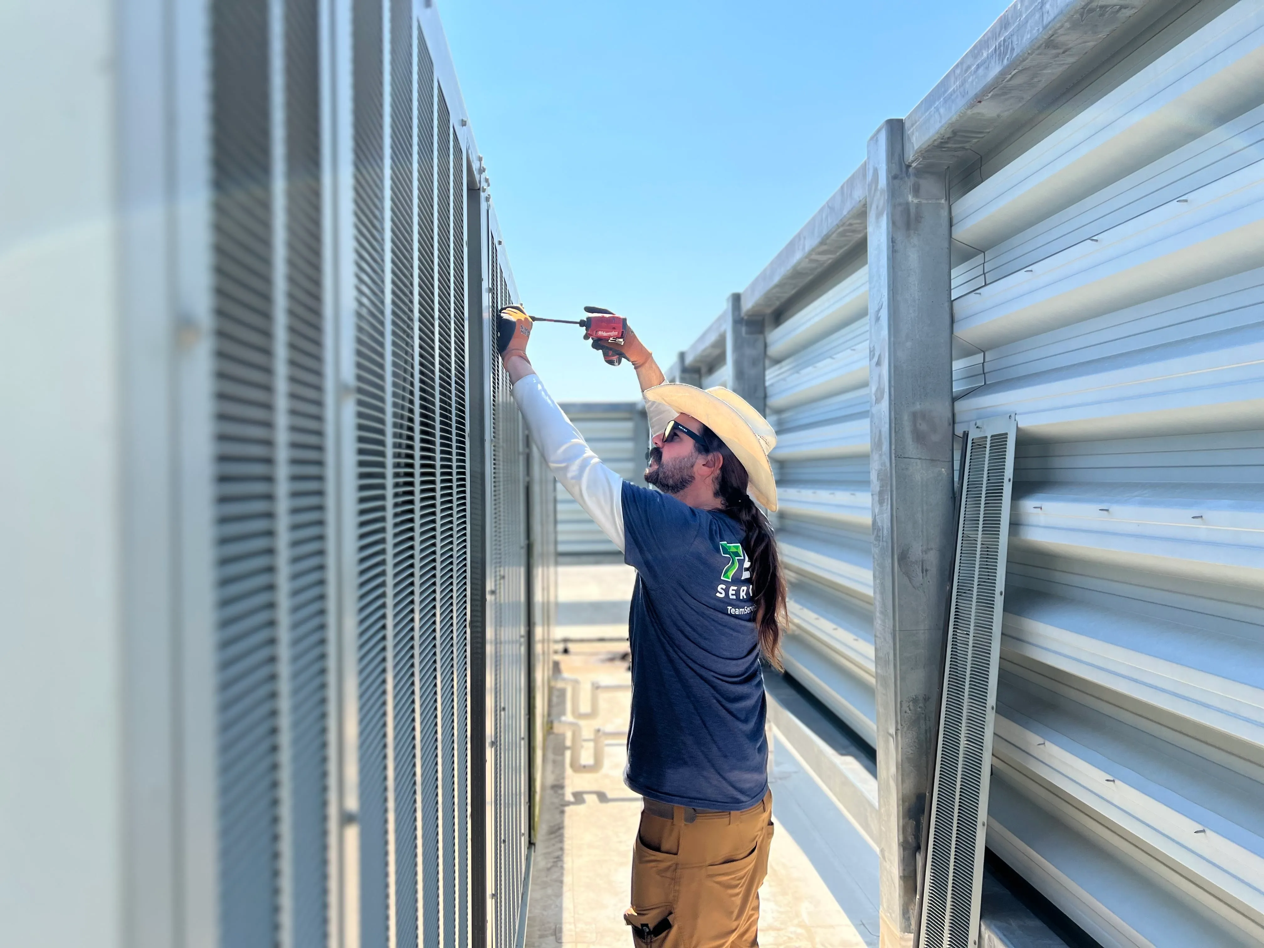 A man wearing sunglasses, a wide-brimmed hat, and work gloves is using a red drill to work on industrial equipment outdoors between metal structures under a clear blue sky.