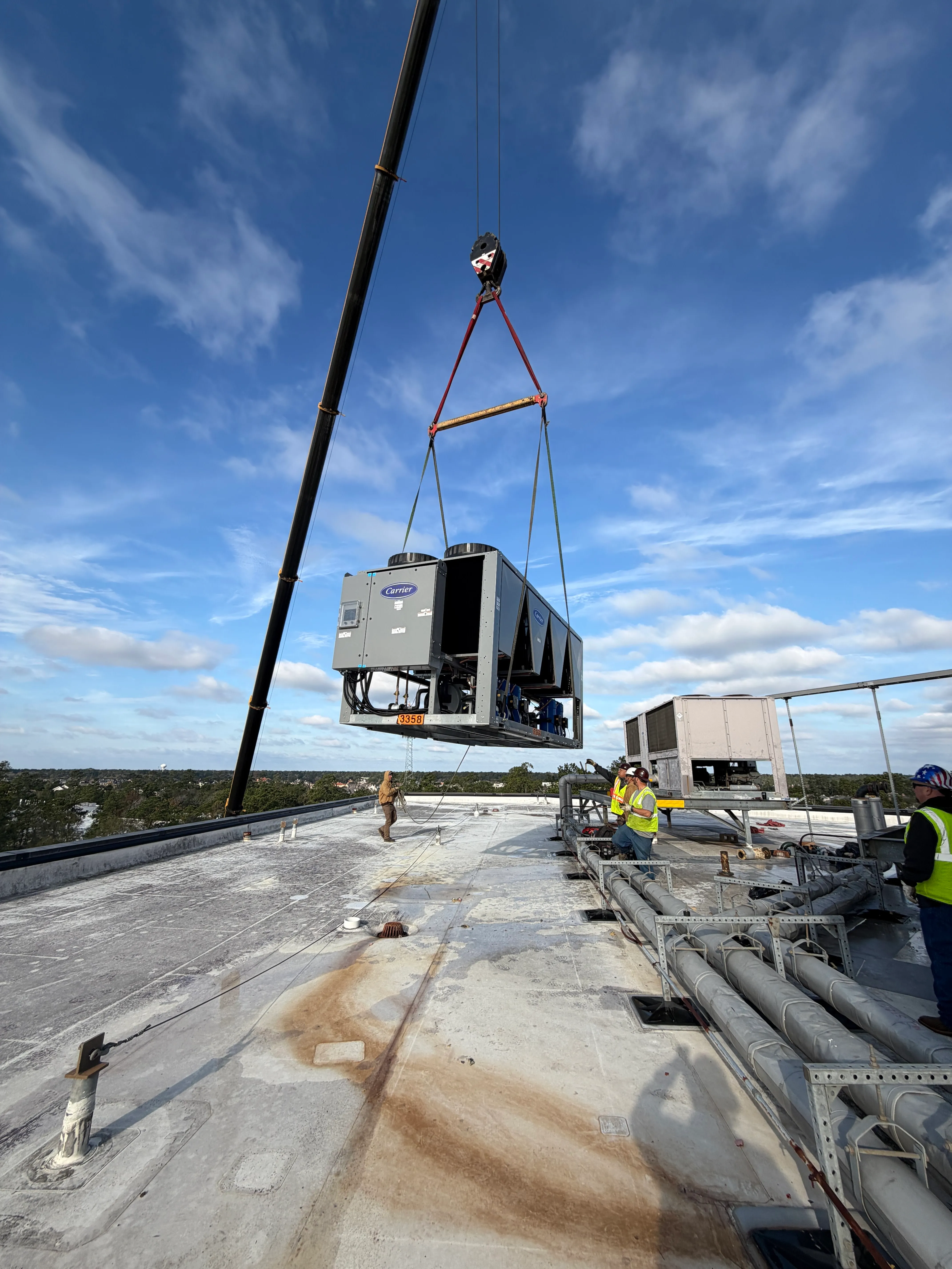 A large HVAC unit with Carrier branding is being lifted by a crane on a flat rooftop where three workers in safety vests and helmets are standing nearby, with a clear blue sky overhead.