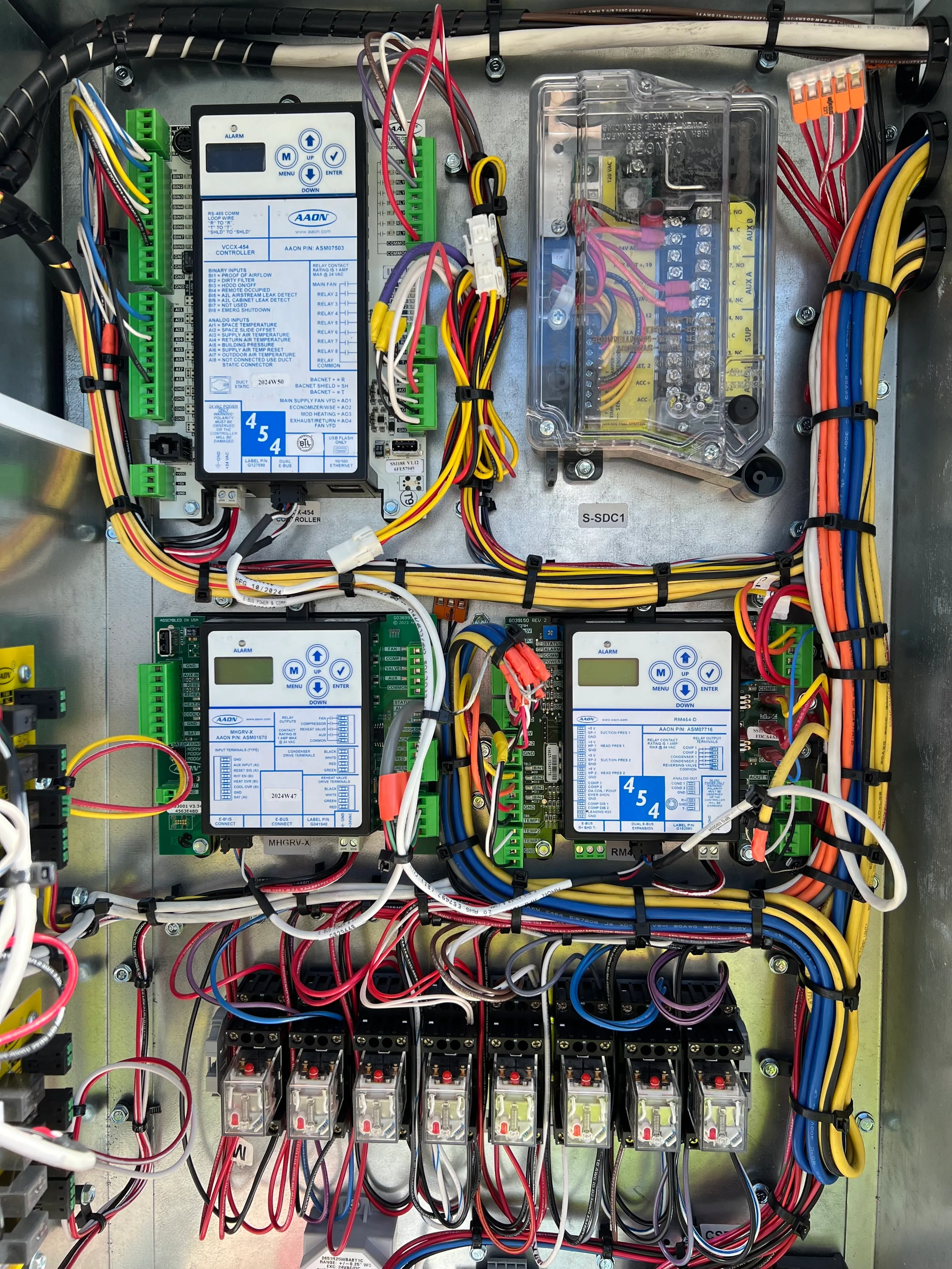 Open electrical control panel with multiple wired circuit boards and relays, neatly organized colored wires bundled with zip ties, and labeled control modules including AAON controllers.