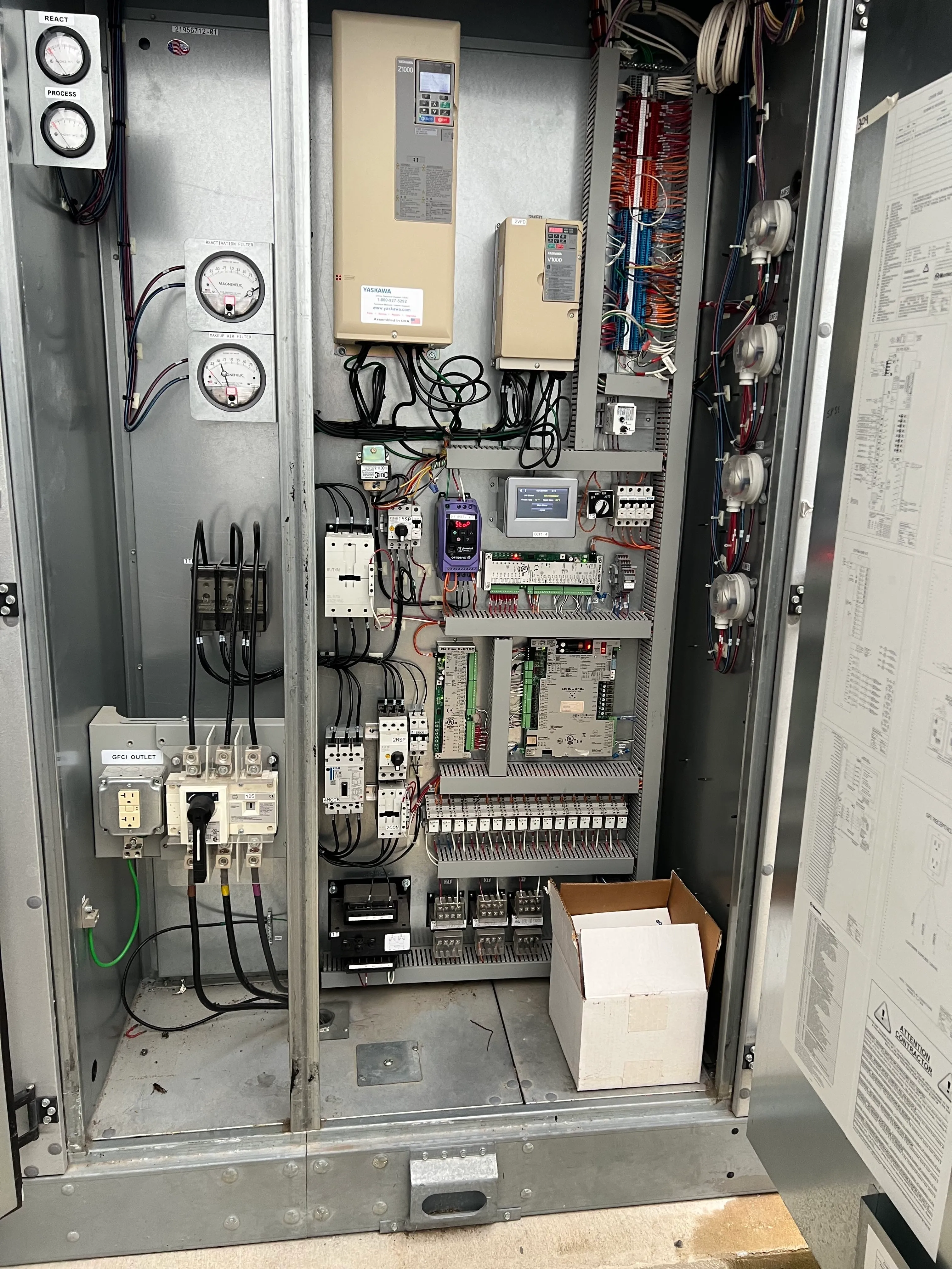 Open industrial electrical control panel cabinet with various components including circuit breakers, wiring terminal blocks, two beige Yaskawa variable frequency drives, gauges labeled React and Process, a purple device displaying 56.0, multiple colored wires organized in bundles, and an open cardboard box on the floor inside the cabinet.