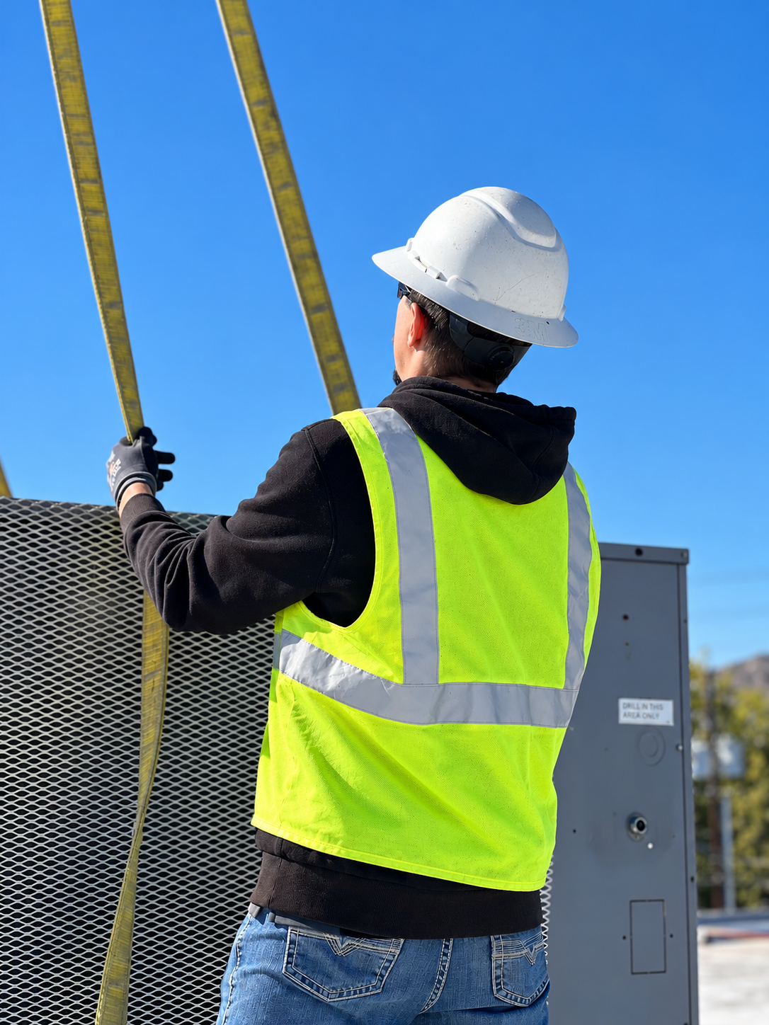 Two construction workers wearing hard hats, safety glasses, and high-visibility vests working on a rooftop HVAC unit, one using a power drill on the unit's panel.