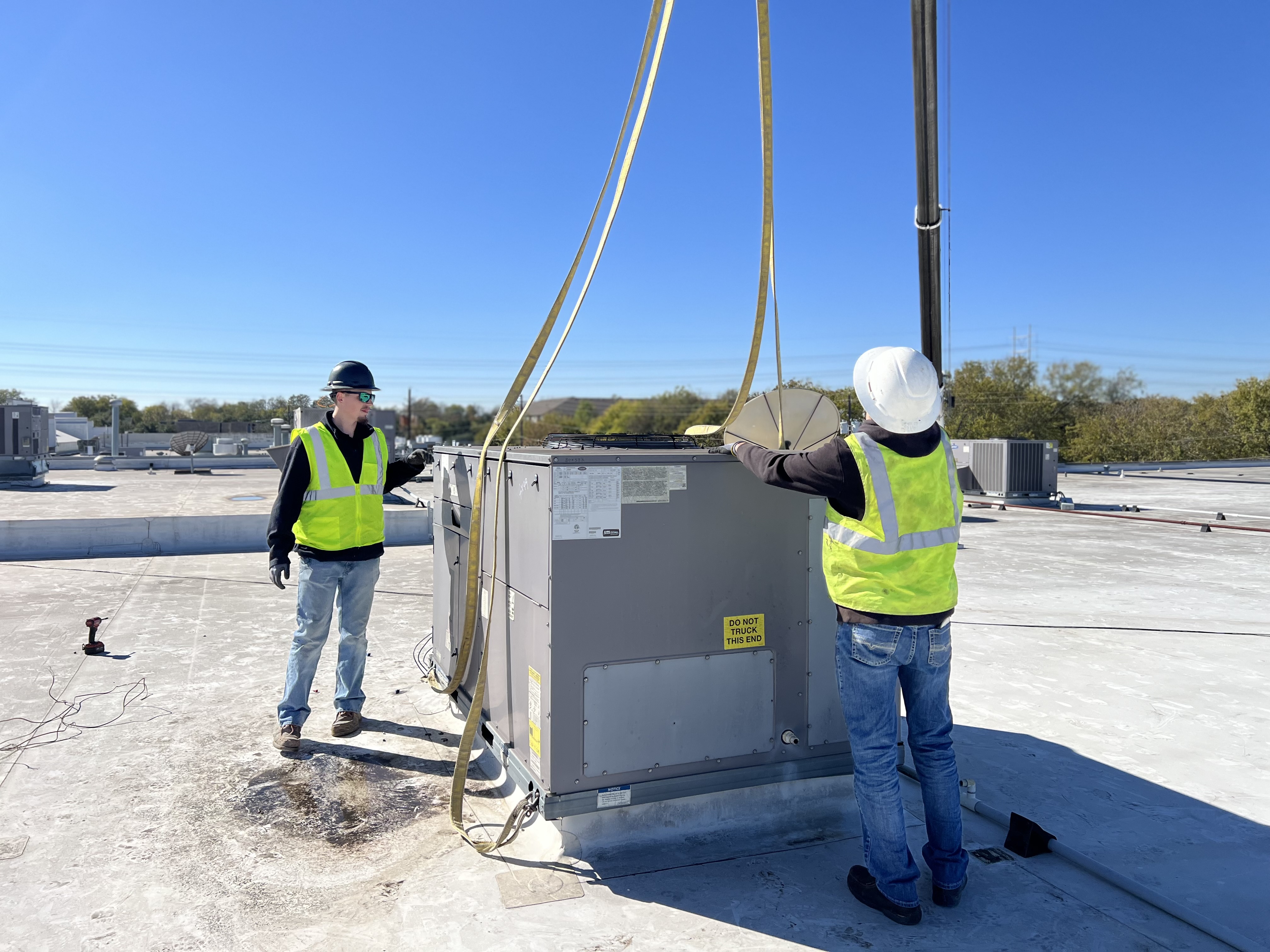 A large HVAC unit with Carrier branding is being lifted by a crane on a flat rooftop where three workers in safety vests and helmets are standing nearby, with a clear blue sky overhead.