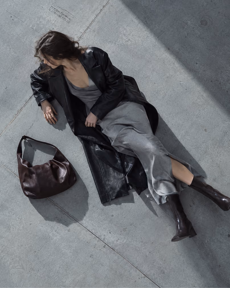 Woman lying on a concrete floor in a black leather coat beside a dark leather bag