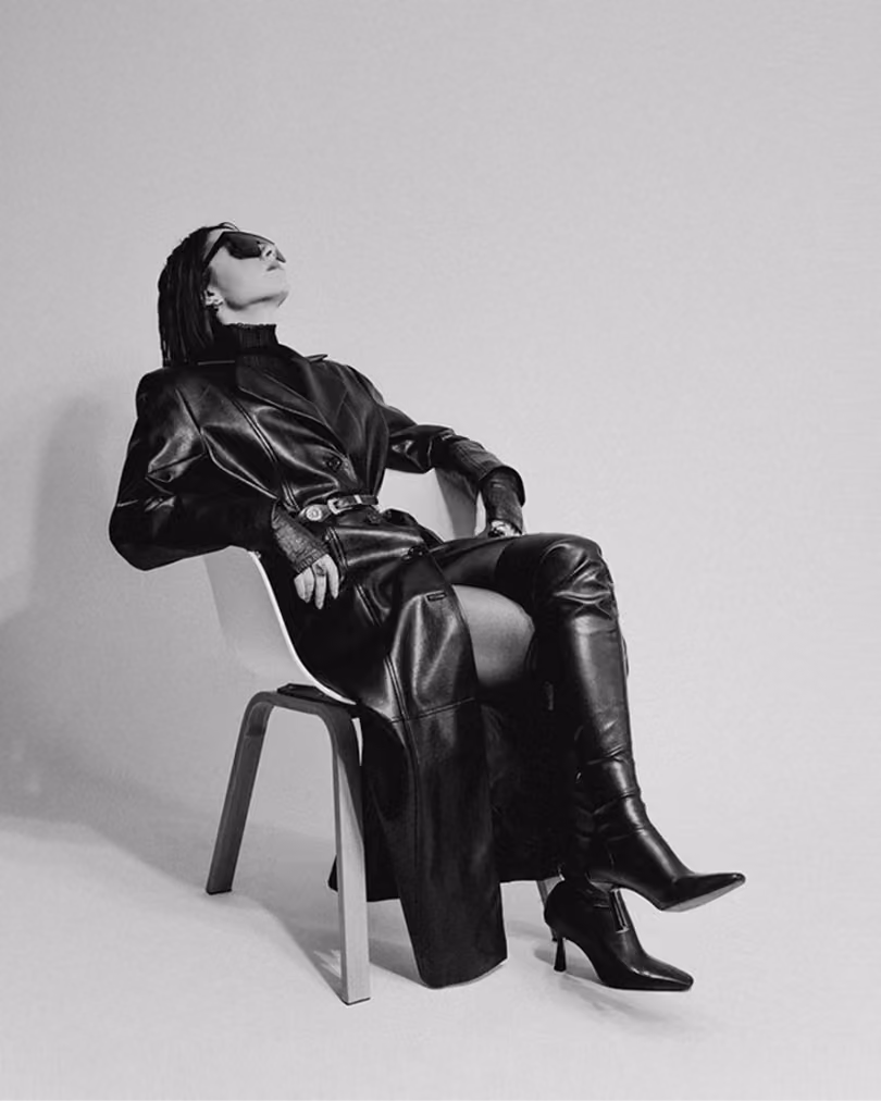 Model seated on a chair in black leather clothing against a pale backdrop