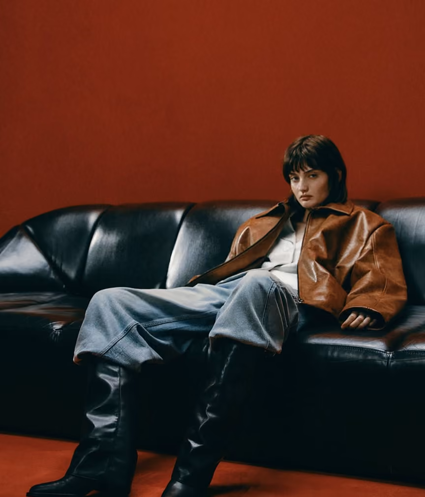 Woman in a tan leather jacket sitting on a black leather sofa against a red wall