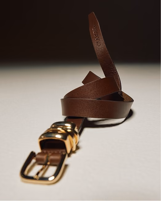 Brown leather belt with a gold buckle coiled on a pale surface