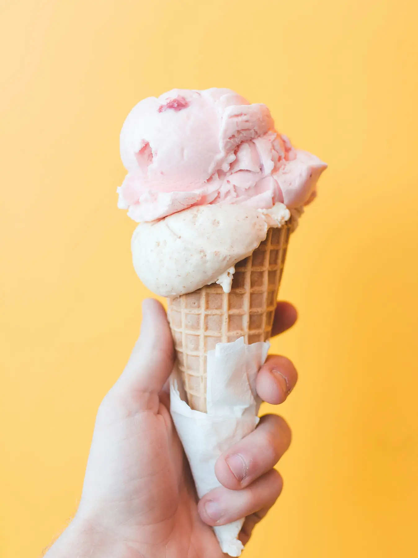 A hand holding a strawberry ice cream cone against a warm yellow ground