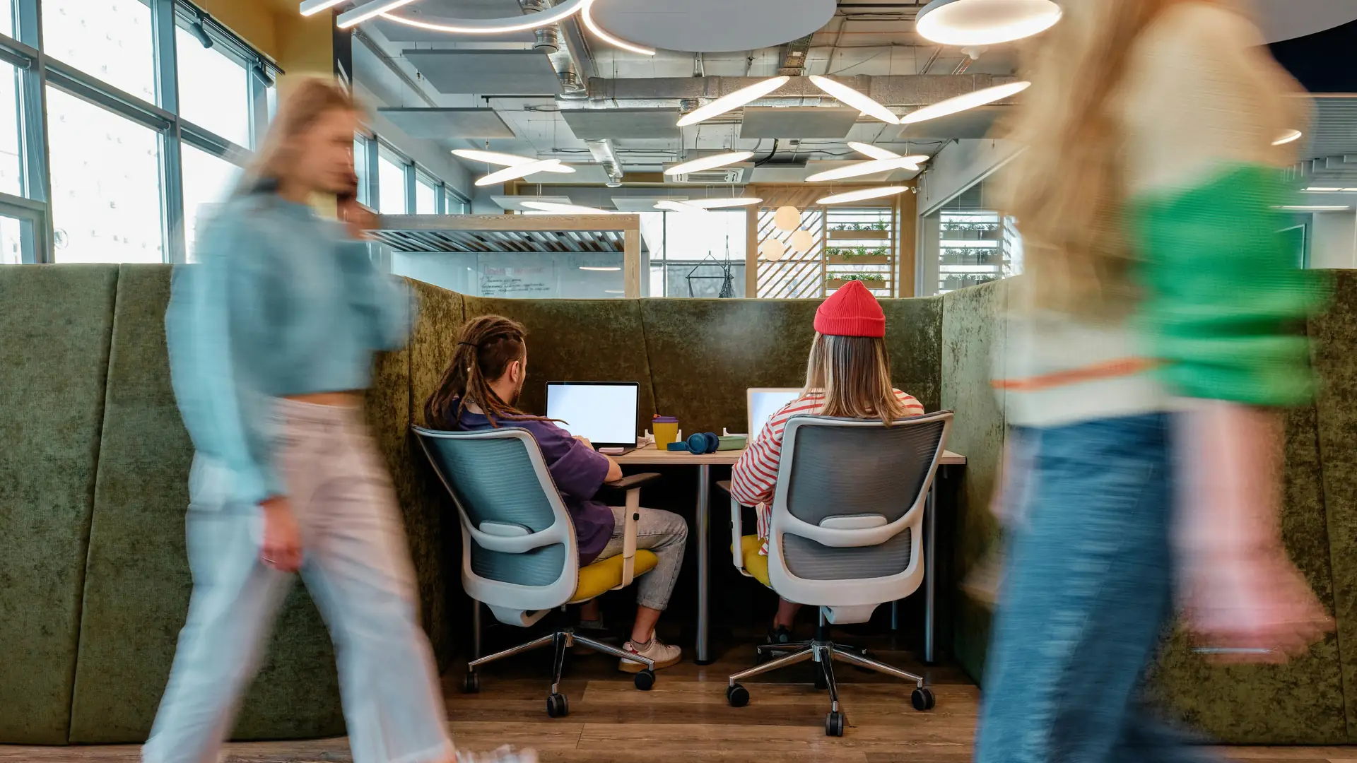 Two people working at laptops in a quiet booth while colleagues walk past in a blur