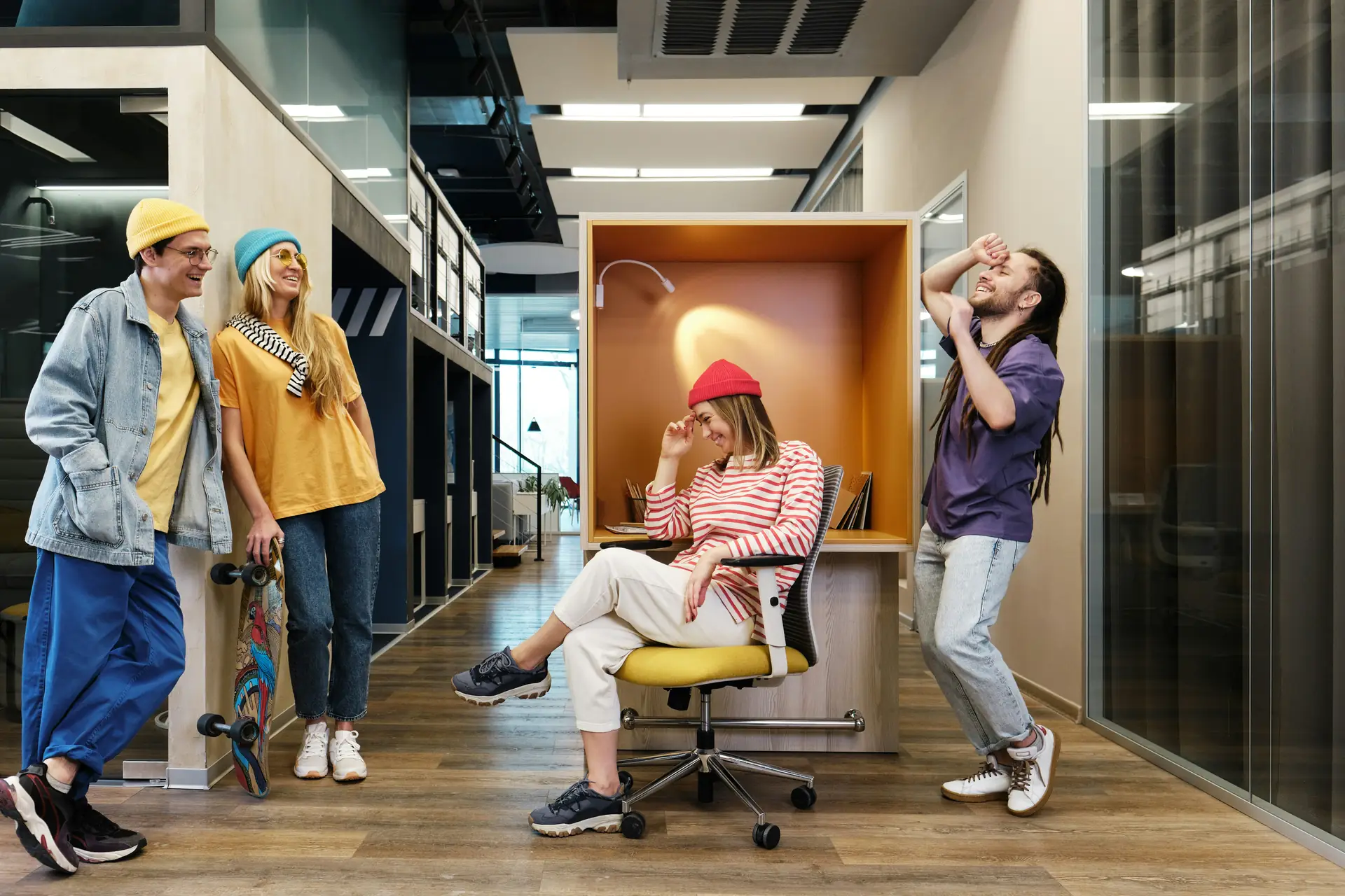 Four colleagues in bright beanies laughing together in an office corridor, one sitting in a work booth